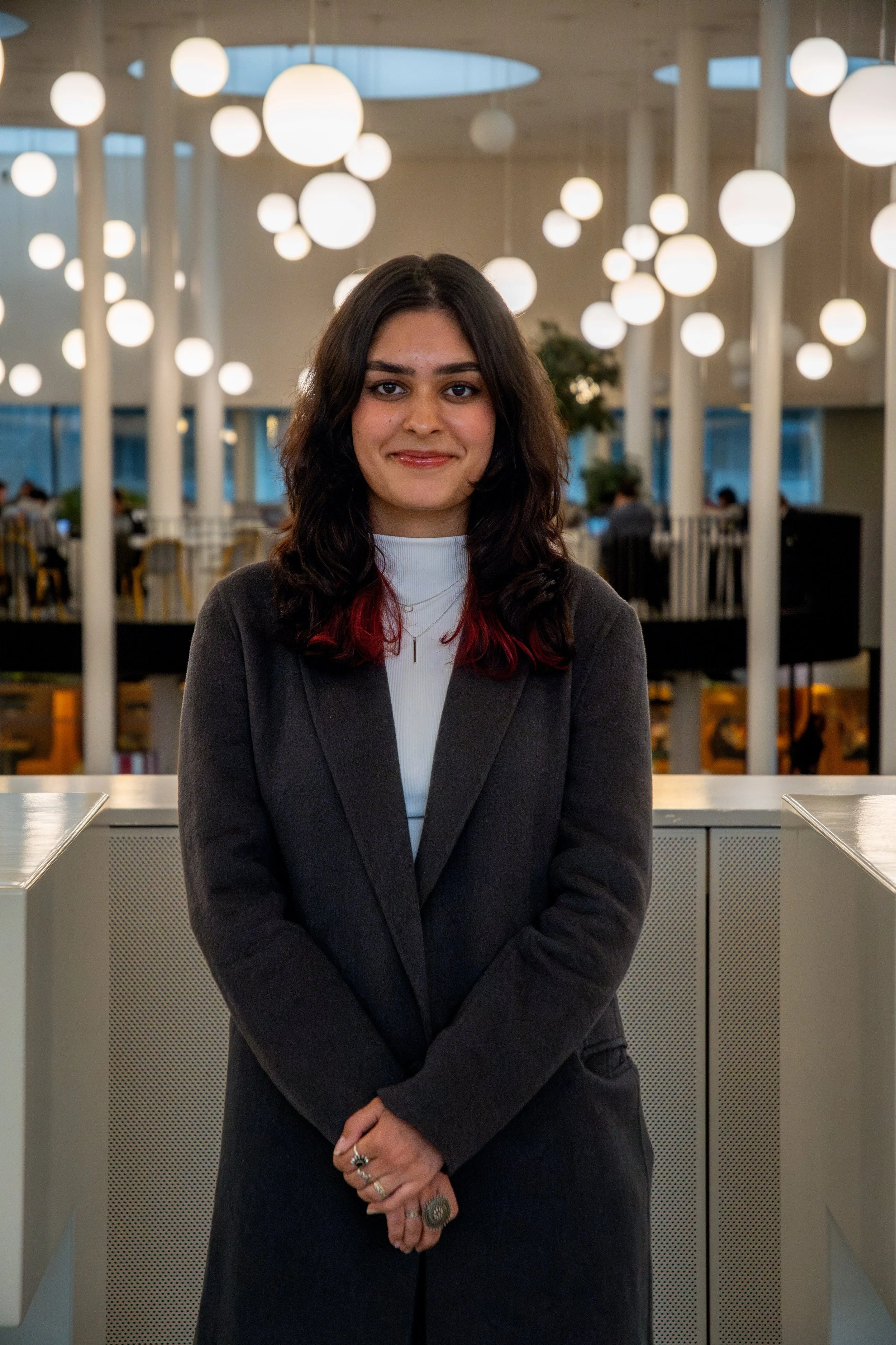 A young female student with dark wavy hair with red tips, wearing a dark gray blazer and white turtleneck, standing indoors with round ceiling lights in the background.