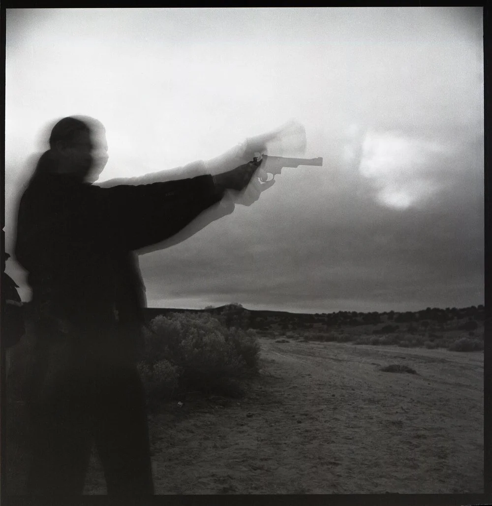 A person aiming a gun, with a motion blur suggesting they are firing, against a cloudy outdoor landscape. Silver Gelatin. Created by Sam Devine Tischler © all rights reserved