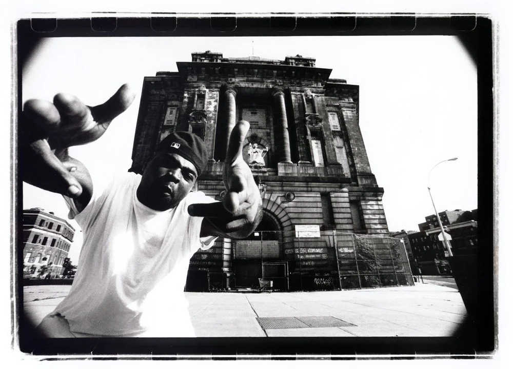 A man wearing a baseball cap and white t-shirt pointing towards the camera in front of a large historic building, with a city street and buildings visible in the background, in a black and white photograph. Created by Sam Devine Tischler © all rights