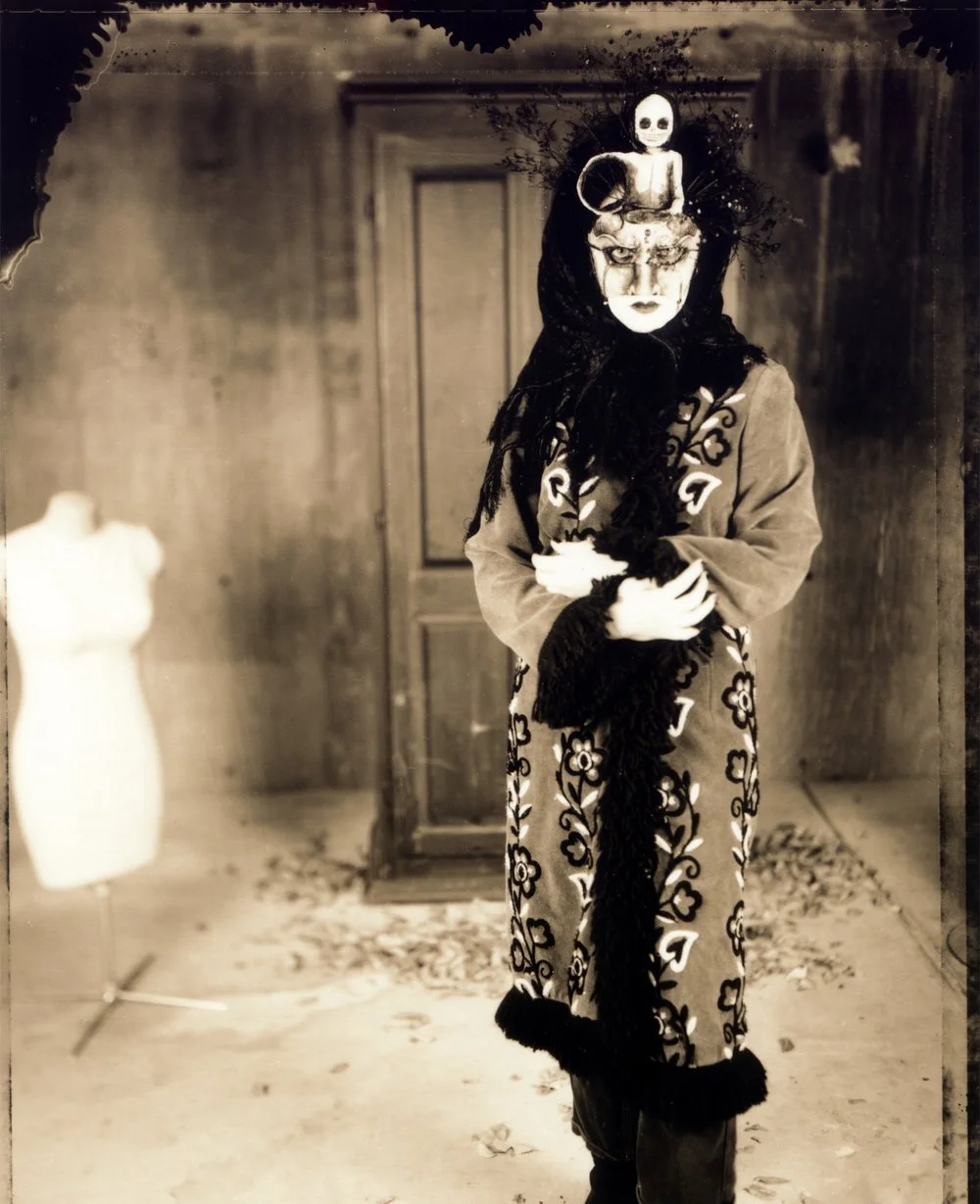 A person in an ornate costume with a mask standing in a vintage room, with a mannequin in the background. Toned silver gelatin. Created by Sam Devine Tischler © all rights reserved