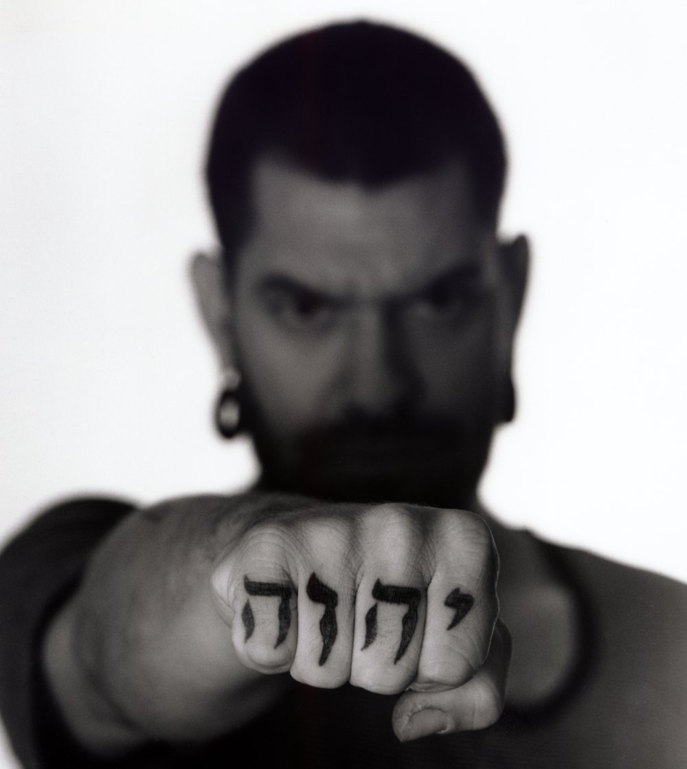 A man with a serious expression points his fist directly at the camera, showing tattoos of Hebrew letters on his fingers, with a blurred background. Created by Sam Devine Tischler © all rights reserved