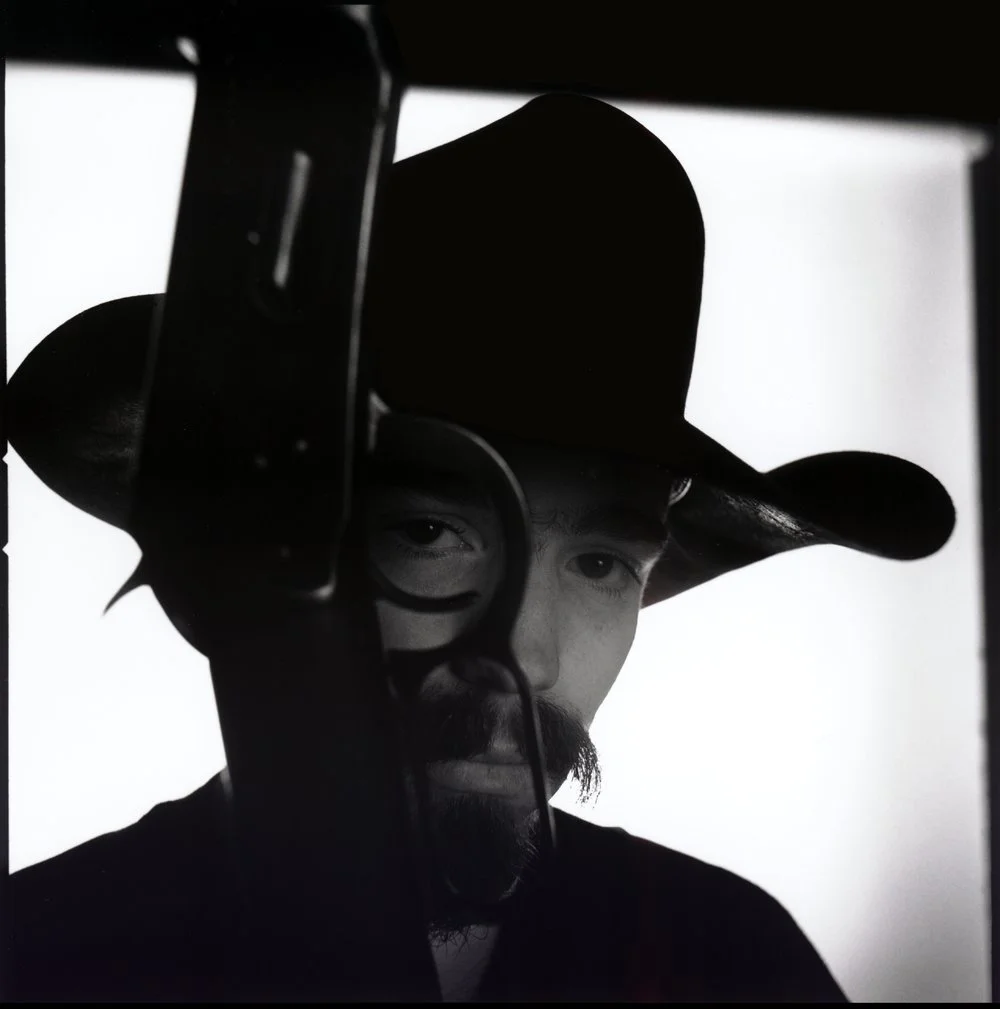 Black and white photo of a man wearing a cowboy hat, looking at the camera through a rifle, with a mustache and beard. Created by Sam Devine Tischler © all rights reserved
