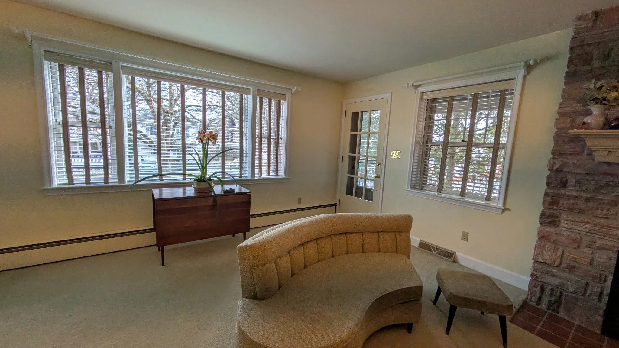 Living room with beige curved sofa, small matching ottoman, wood sideboard with plant, large window with blinds, white door with glass panes, brick fireplace on the right, beige walls, carpeted floor, and a few decorations.
