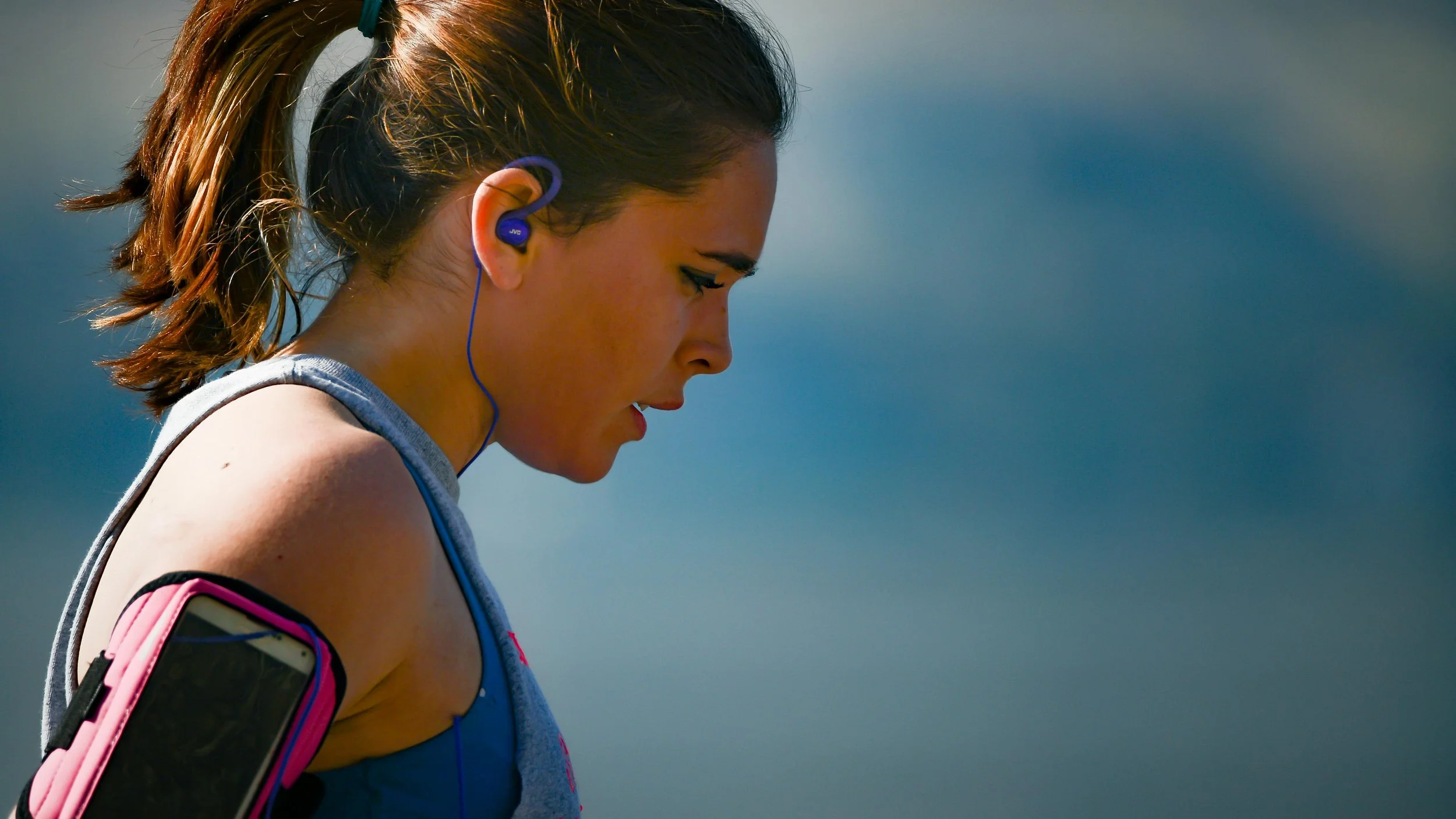 Side view of a young woman during outdoor exercise, wearing earphones, a sleeveless top, and a smartphone armband, with a blurred natural background.