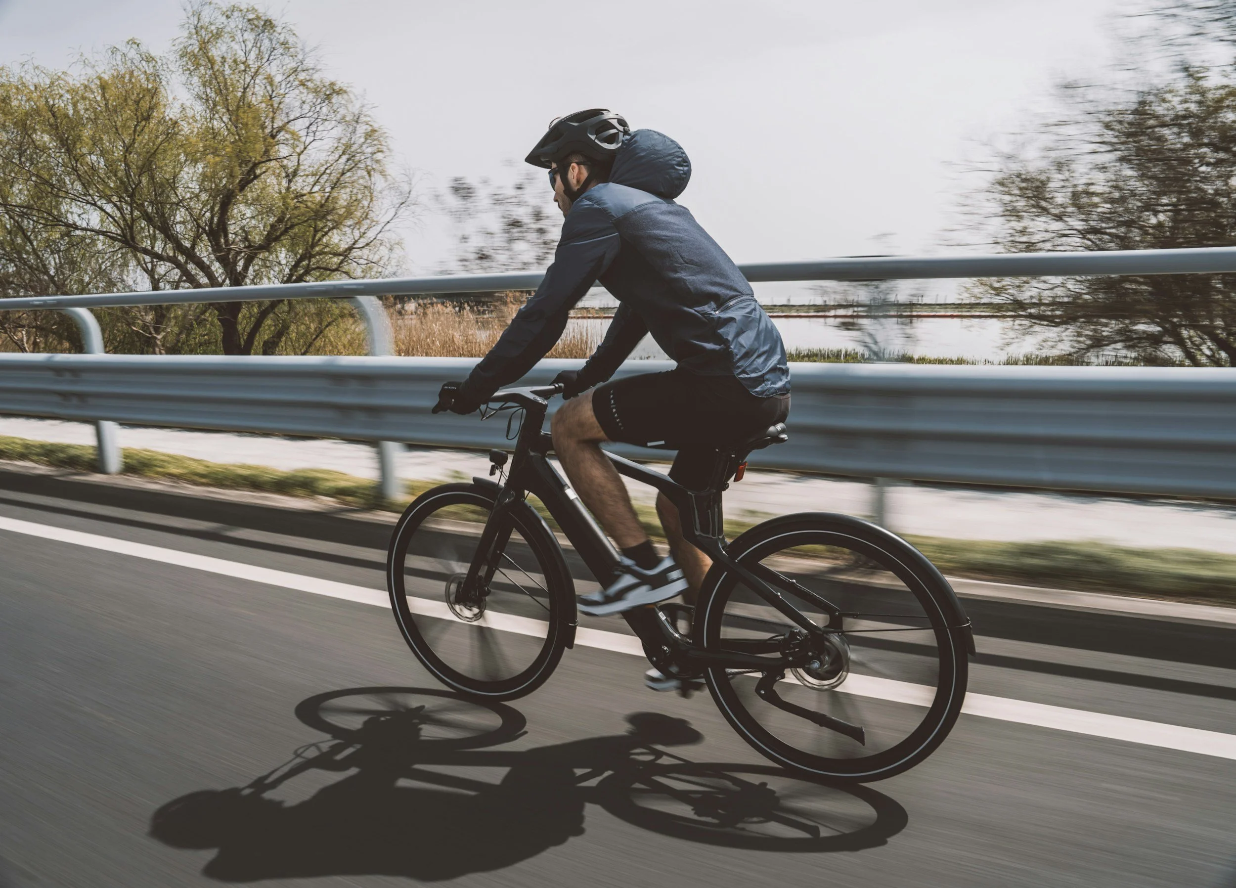 A person riding a black electric bicycle on a paved road, wearing a black helmet, dark jacket, black shorts, and sneakers, with trees and a metal guardrail in the background.