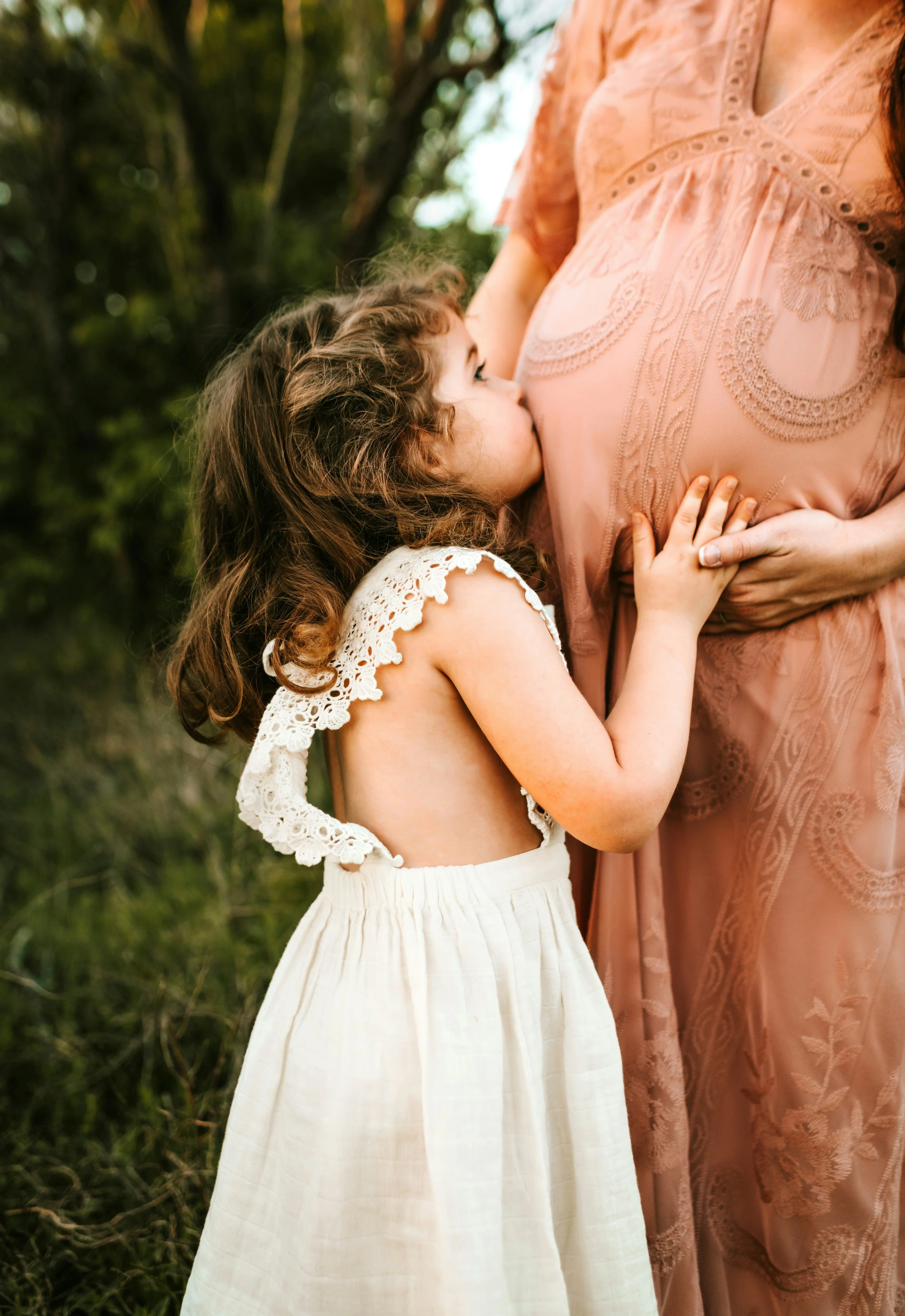 A young girl with curly brown hair, wearing a white dress with lace details, is kissing the pregnant belly of a woman dressed in a peach-colored embroidered dress on a nature backdrop.