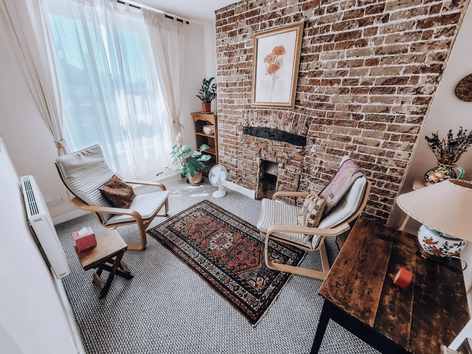 Cozy living room with exposed brick fireplace, two armchairs, a small side table with tissue box, potted plants, wooden shelves, and a colorful rug, with large window drapes letting in natural light.