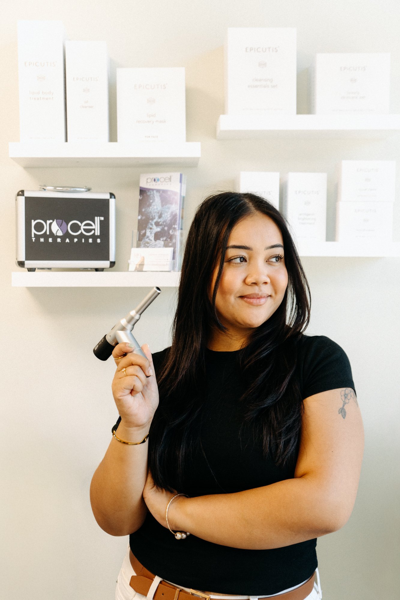 A woman with long dark hair holding a medical device, standing in front of shelves with skincare products at Procet Therapies.