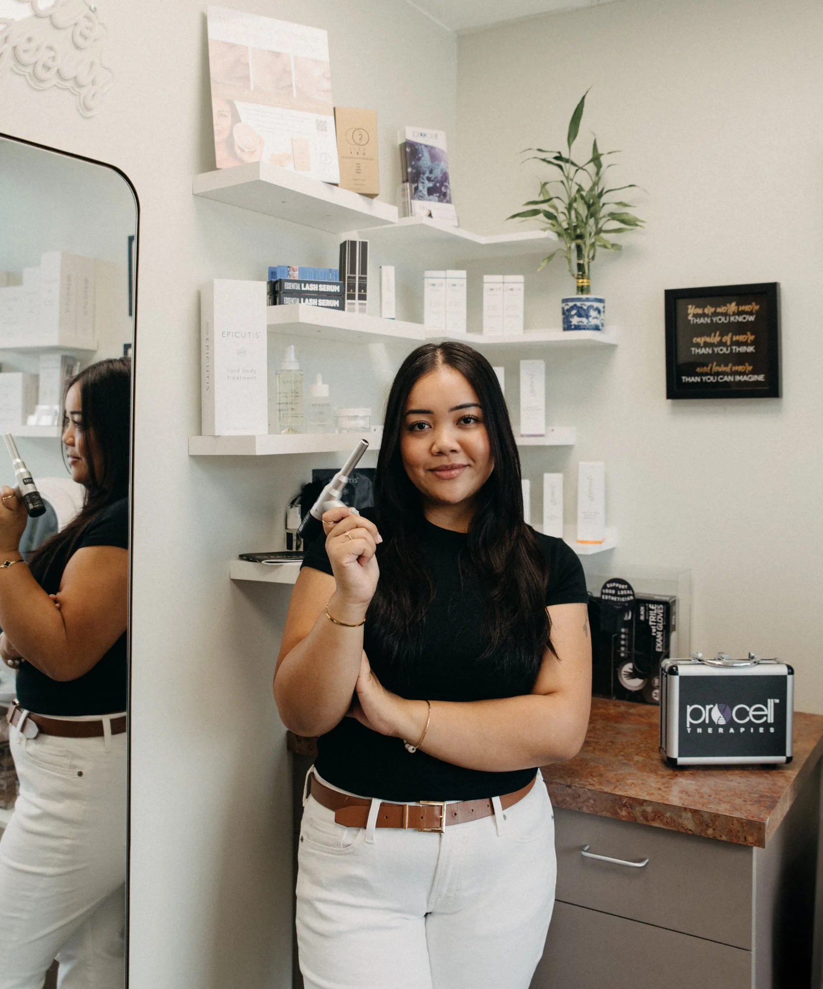 A woman with long dark hair holding a medical device, standing in front of shelves with skincare products at Procet Therapies.