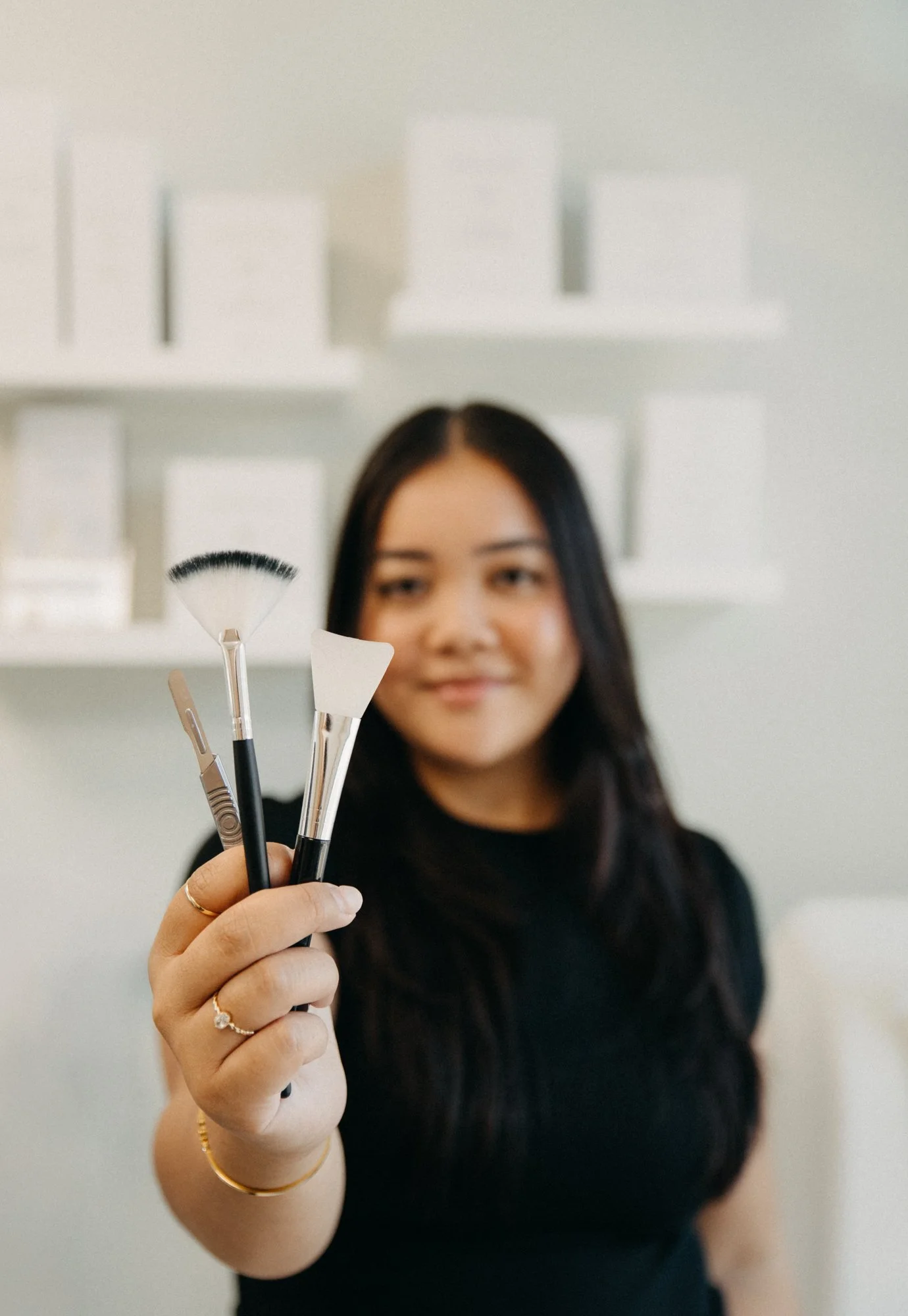 A woman in a black shirt holding up three makeup tools, including an eyebrow brush, a spatula, and a small fan brush, with a blurred background of white shelves and boxes.