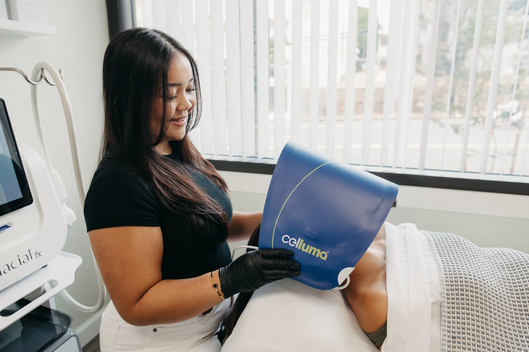 A woman with long dark hair, wearing a black shirt and black gloves, smiling while holding a blue medical device labeled 'celluma' near a patient's neck in a clinical setting with bright natural light coming through window blinds.