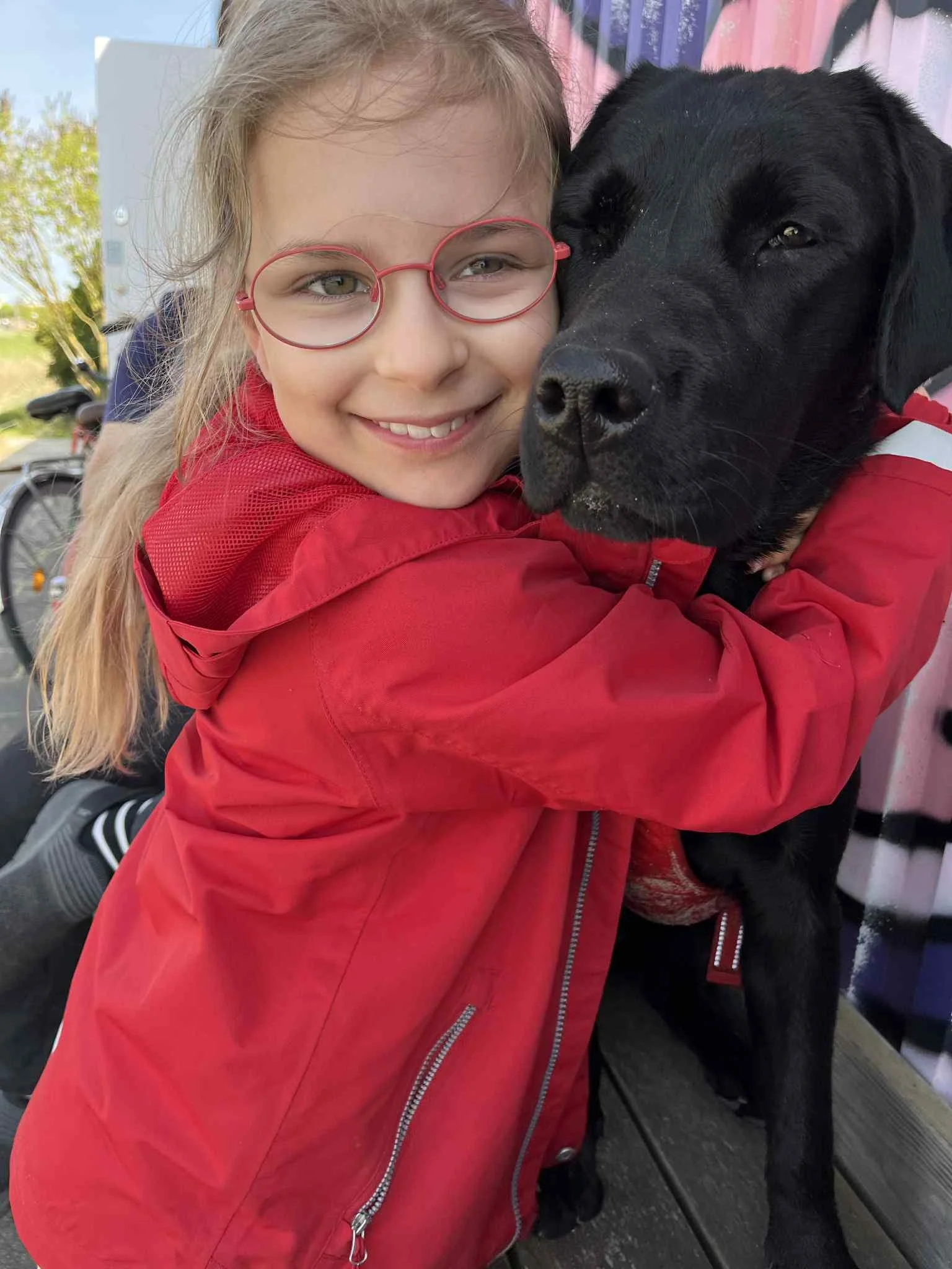 A young girl with glasses and a red jacket hugging a black dog outdoors.