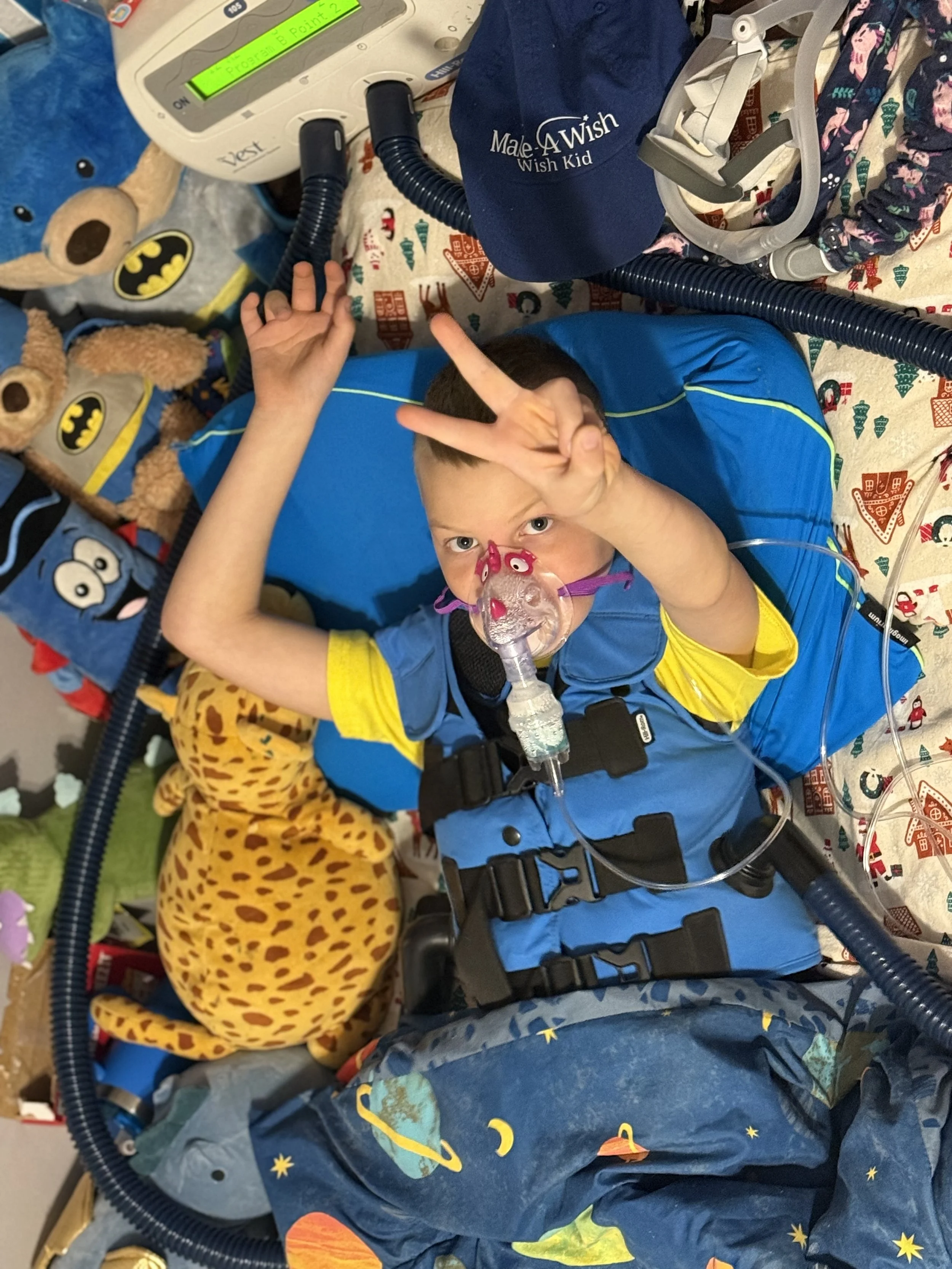 Child lying in hospital bed with a breathing mask, making a peace sign with hand, surrounded by teddy bears, medical equipment, and festive blankets.