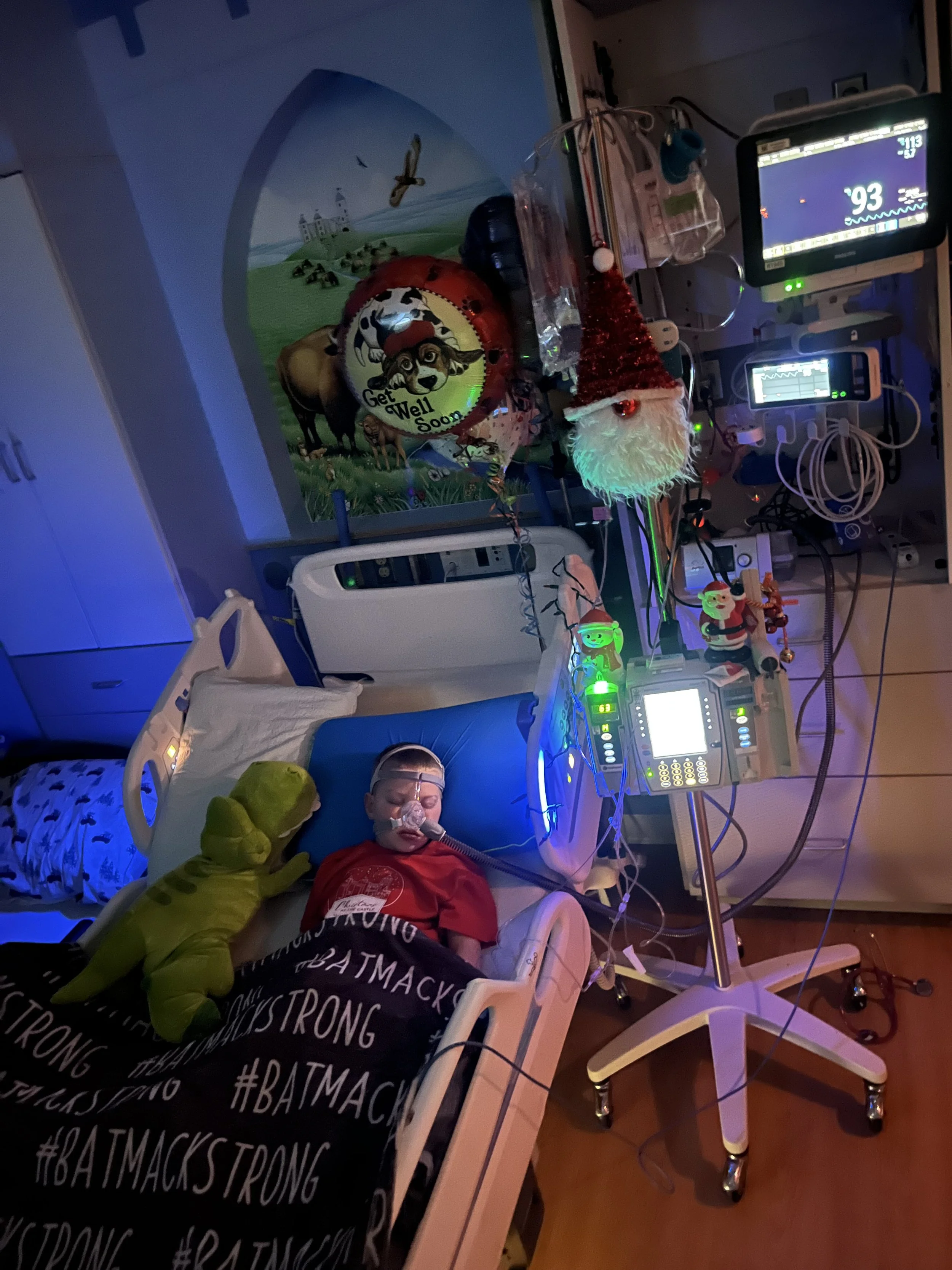 A young child in a hospital bed wearing an oxygen mask, with a green dinosaur plush toy. The bed is covered with a black blanket with white hashtag text '#BATMACSSTRONG'. The room is decorated with Christmas and holiday items, including a Santa elf hat, a Santa figurine, and balloons with festive messages. Medical equipment and monitors are visible next to the child.