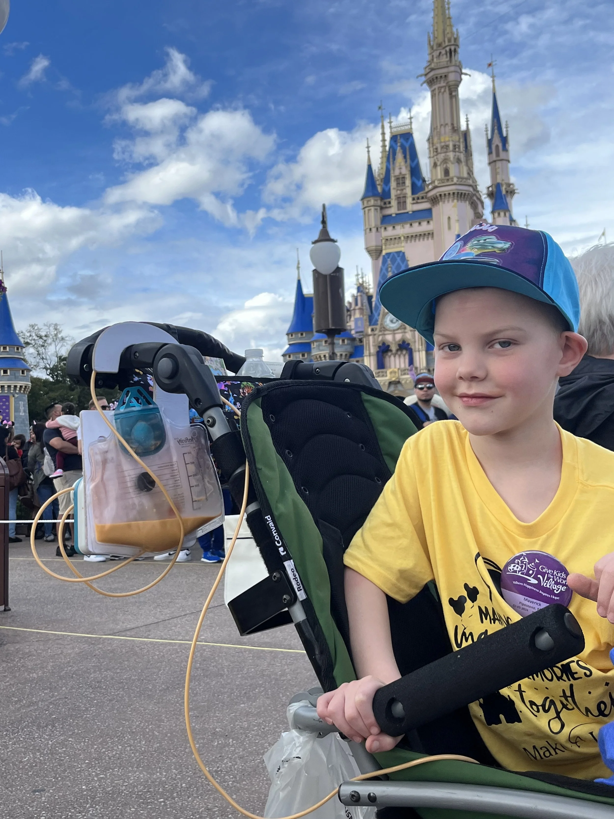 A young boy sitting in a stroller, wearing a blue cap and yellow shirt, at Disneyland with Cinderella's Castle in the background. There is a medical IV bag hanging from the stroller.