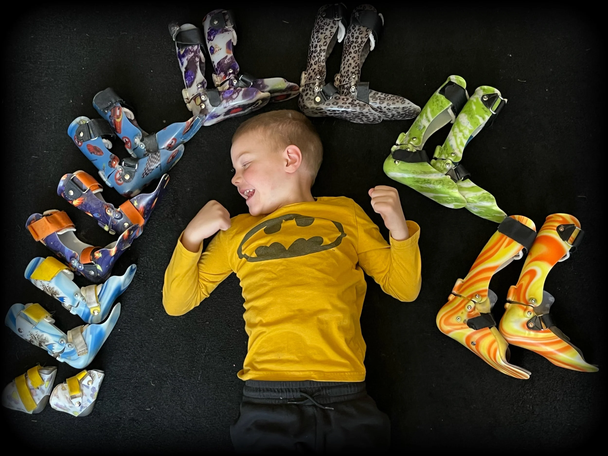 A smiling young boy lying on a black carpet, wearing a yellow Batman t-shirt, surrounded by a colorful arrangement of ski boots forming the word 'LOVE'.