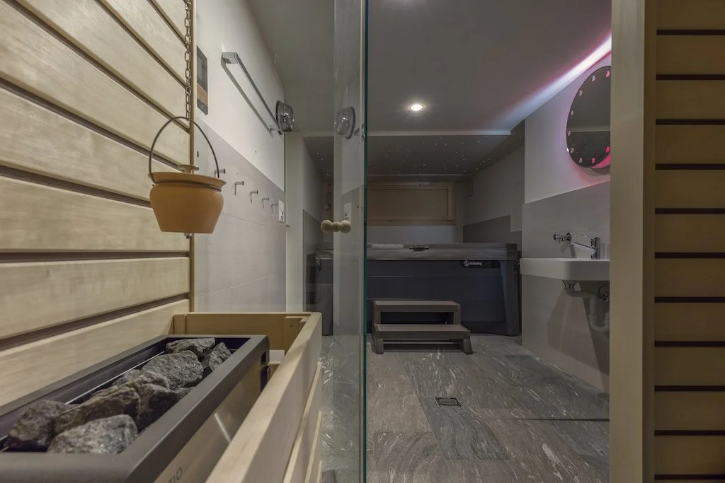 Innenraum eines Sauna- und Wellnessbereichs mit Holzelementen, Sauna, Whirlpool und Waschbecken, dekorative Beleuchtung.