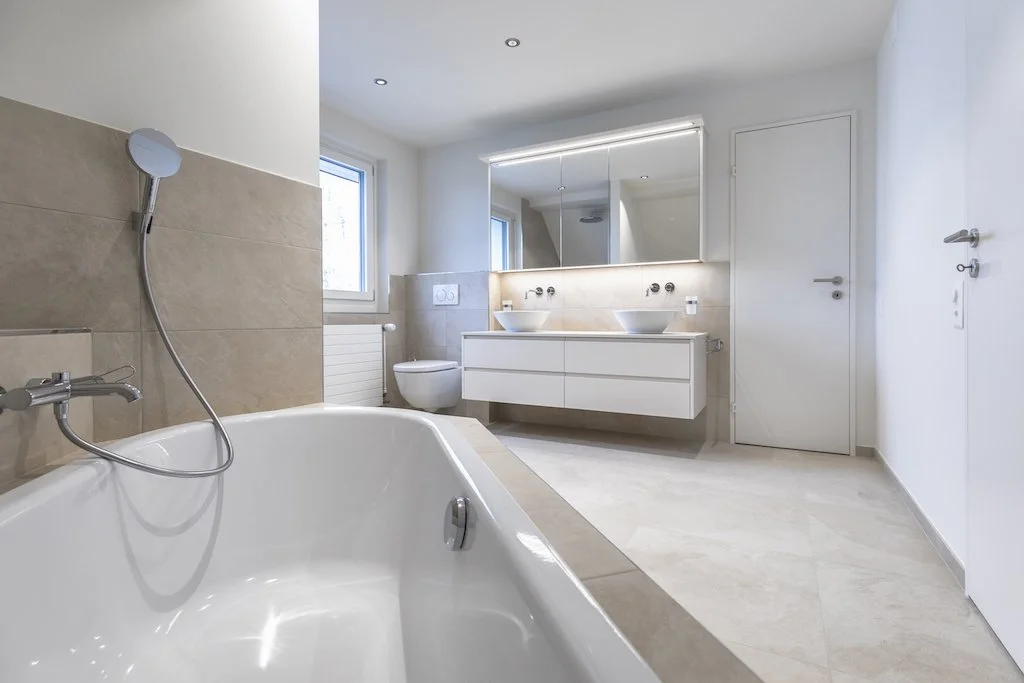 Moderner Badezimmer mit Badewanne, Doppelwaschbecken, wandhängendem WC, Spiegel und hellem Fliesenboden.