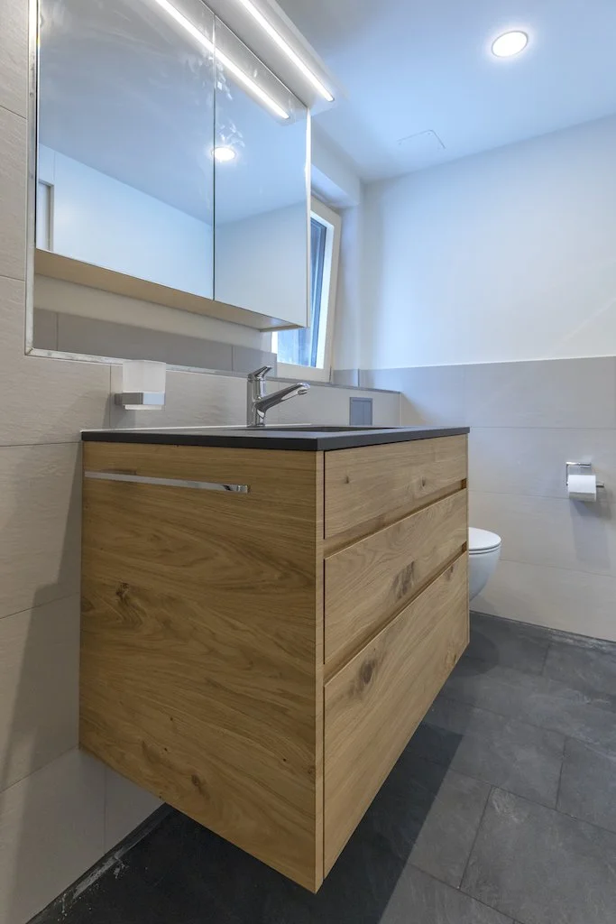 Modernes Badezimmer mit Holz-Waschtisch, Spiegel mit Beleuchtung, Fenster und Toilette.