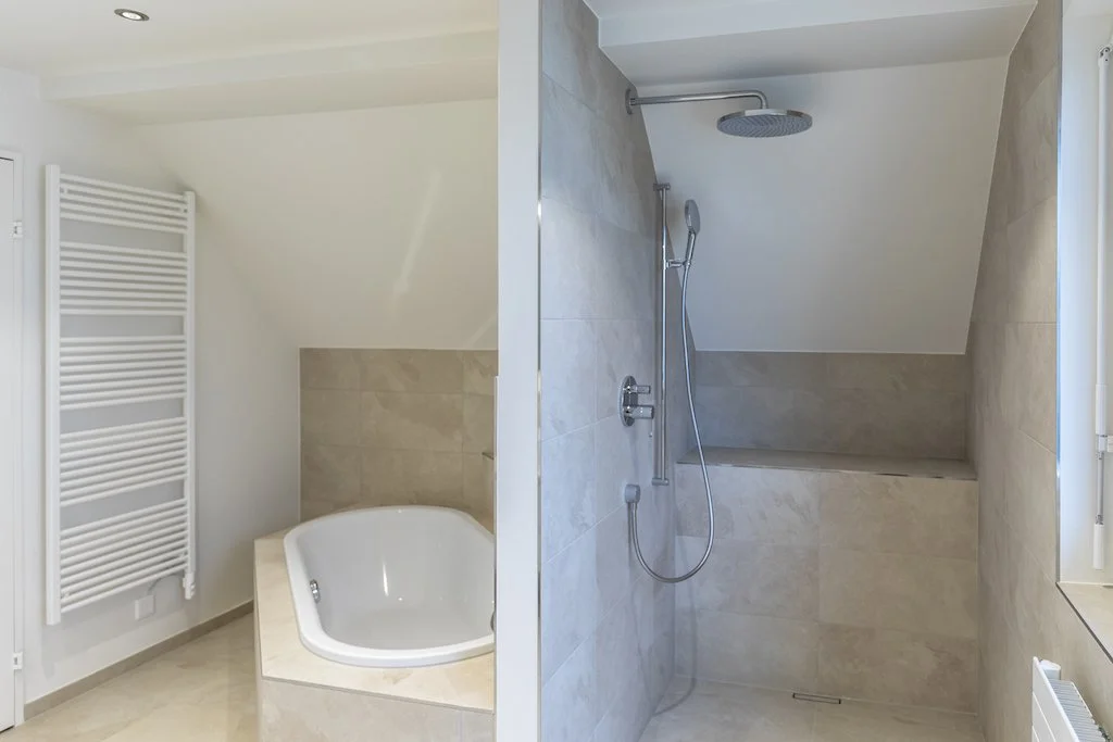 Badezimmer mit Jacuzzi und Dusche, getrennt durch eine Wand, mit beige Fliesen und Wandheizkörper.