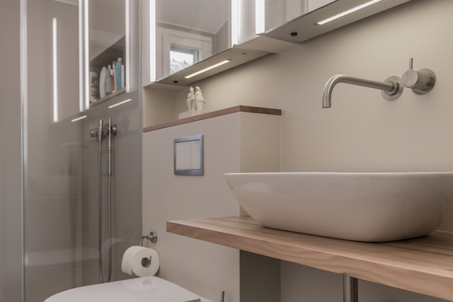 Modernes Badezimmer mit einer weißen Waschschüssel auf einem Holz-Waschtisch, einem beleuchteten Spiegelschrank, einem Handtuchring mit Toilettenpapier, einer Dusche mit Glastür und einer kleinen Fensterseite.