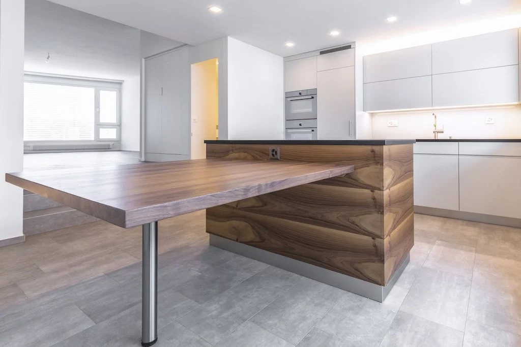 Moderne, helle Küche mit weißem Schrank, Holzarbeitsfläche und einem großen Holzblock als Theke oder Esstisch, lichtdurchflutet mit großer Fensterfront im Hintergrund.