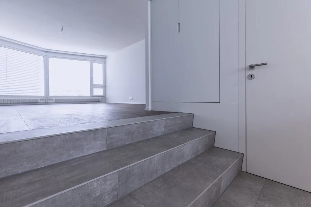 Innenraum mit Treppen, weiße Wände, große Fenster mit Jalousien, gefliester Boden, geschlossene weiße Tür.