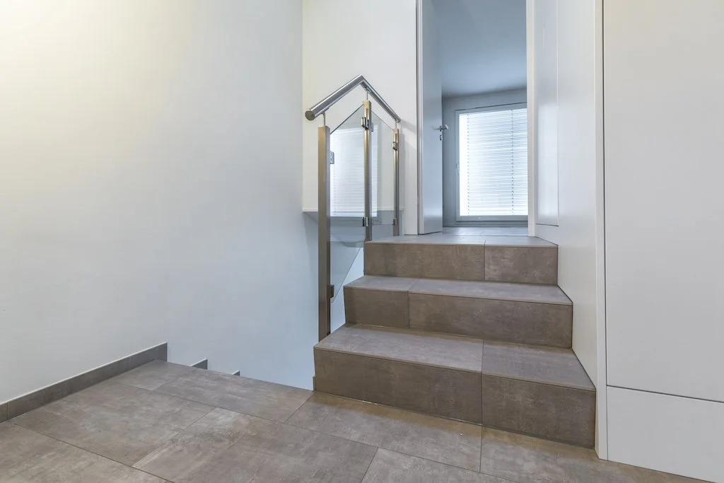 Innenansicht eines Treppenhauses mit drei Stufen, einem Handlauf aus Metall und einem Fenster im Hintergrund.
