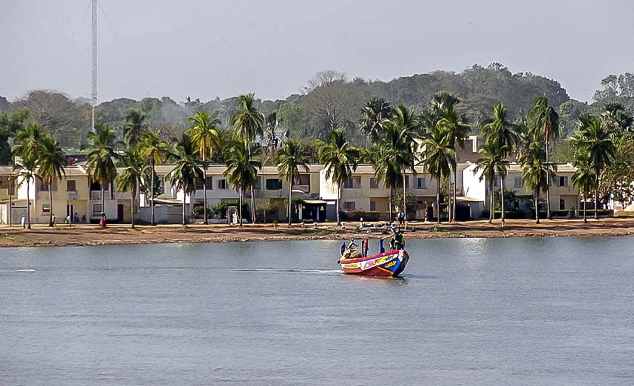 Bateau naviguant sur une rivière avec un front de maisons et des palmiers en arrière-plan.