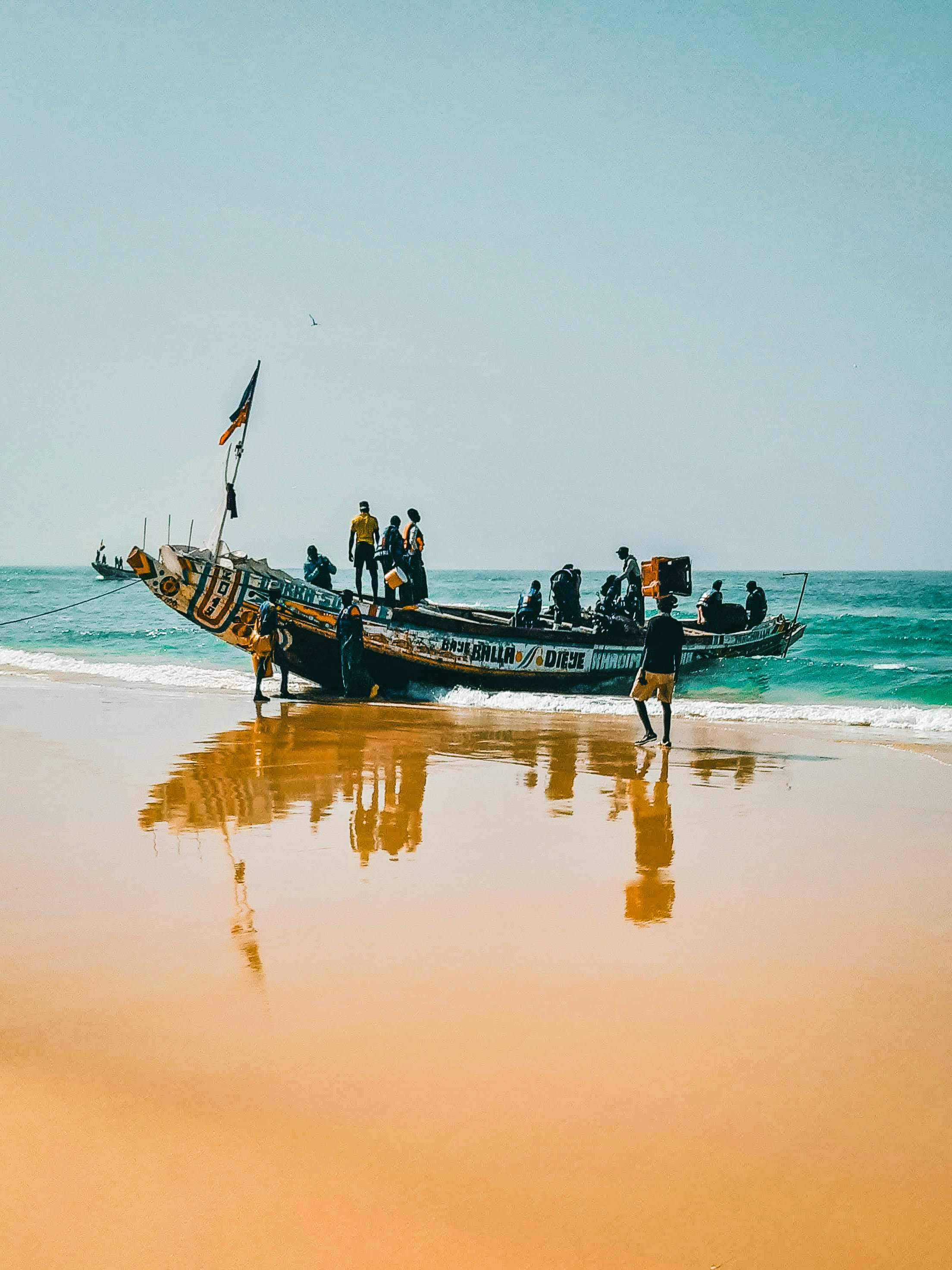 Un bateau traditionnel avec plusieurs personnes à bord arrive sur une plage en sable. La mer est calme et le ciel est clair.