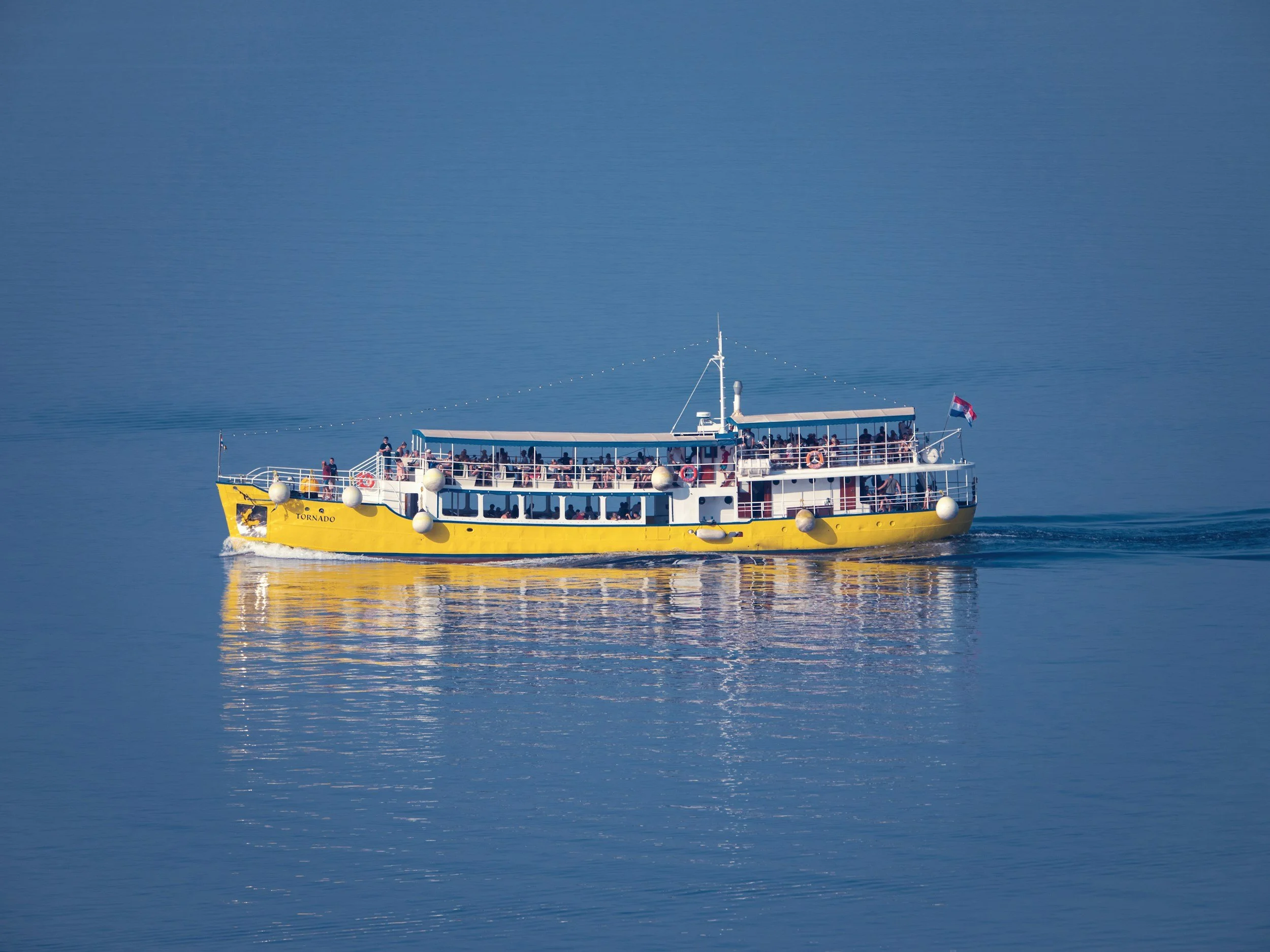 Un bateau jaune nommé Tornado navigue sur une mer calme, avec des passagers à bord, sous un ciel clair.