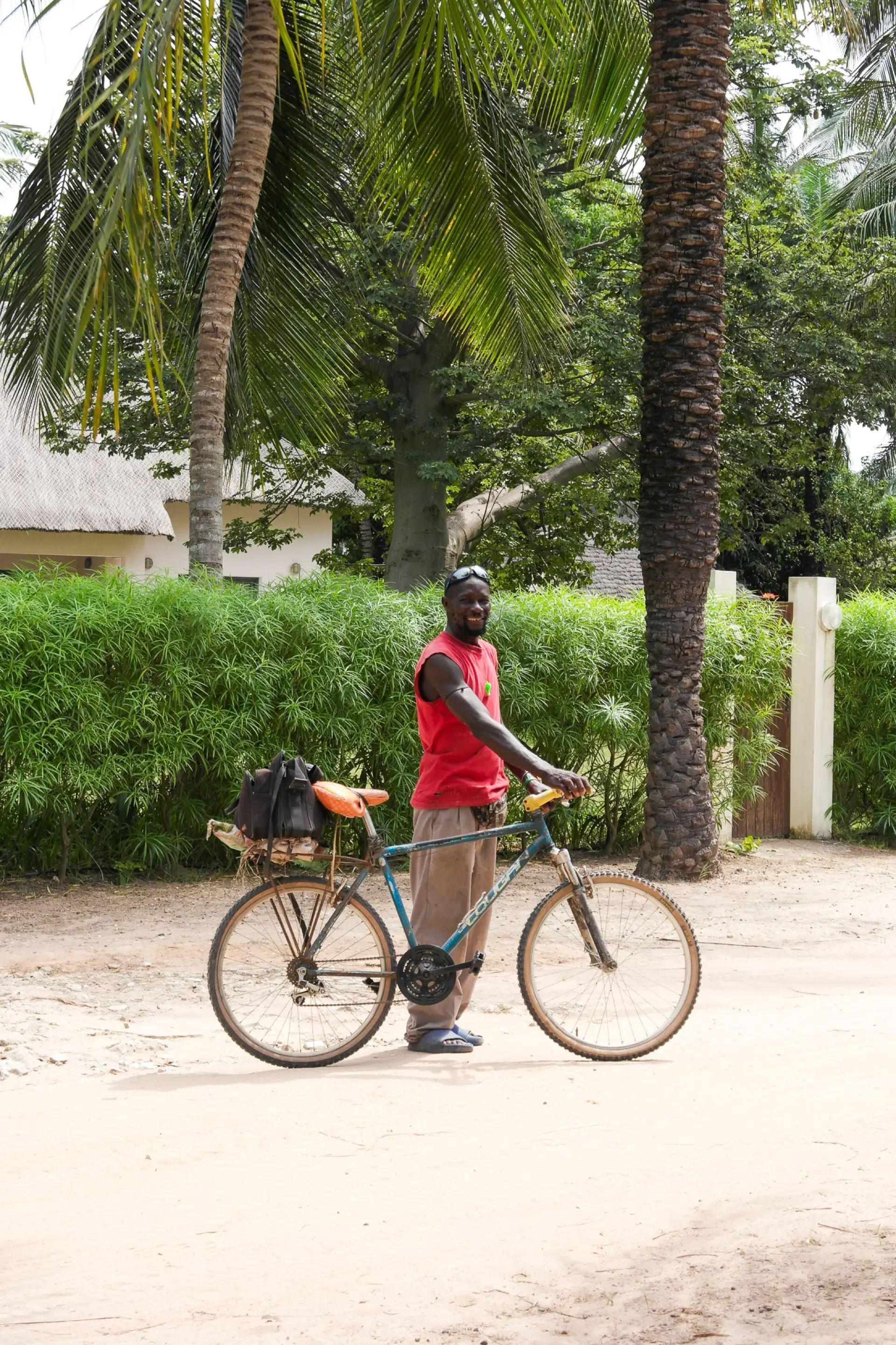 Un homme souriant avec un vélo, portant un t-shirt rouge, dans un environnement tropical avec des palmiers et des arbustes