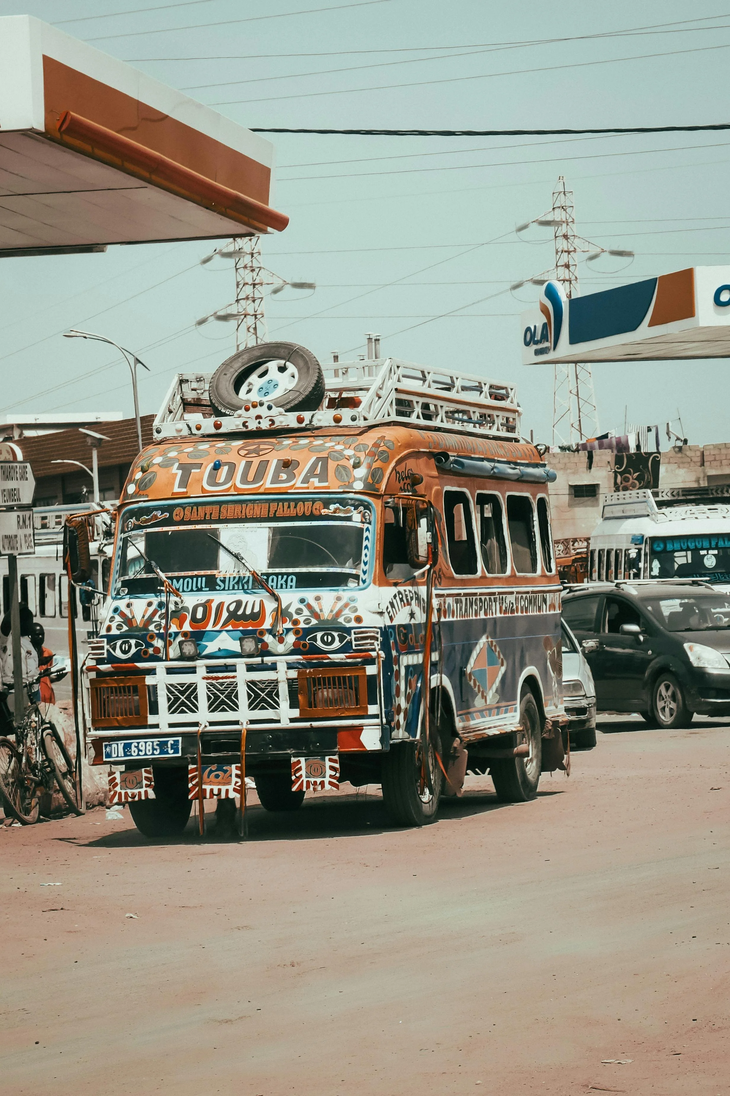 Bus coloré et décoré avec des motifs artistiques, stationné sur une route en zone urbaine avec autres voitures et panneaux publicitaires.