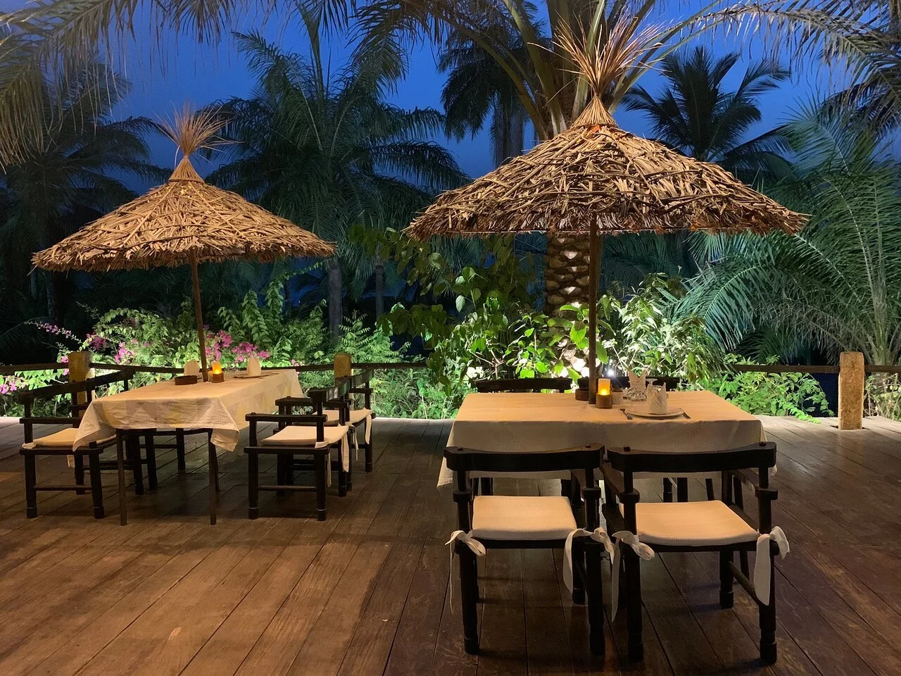 Terrasse de restaurant avec deux tables couvertes de nappes blanches, chaises en bois avec coussins, parasols en paille, bougies allumées, entourée de végétation tropicale avec des palmiers et des plantes. Ciel nocturne.