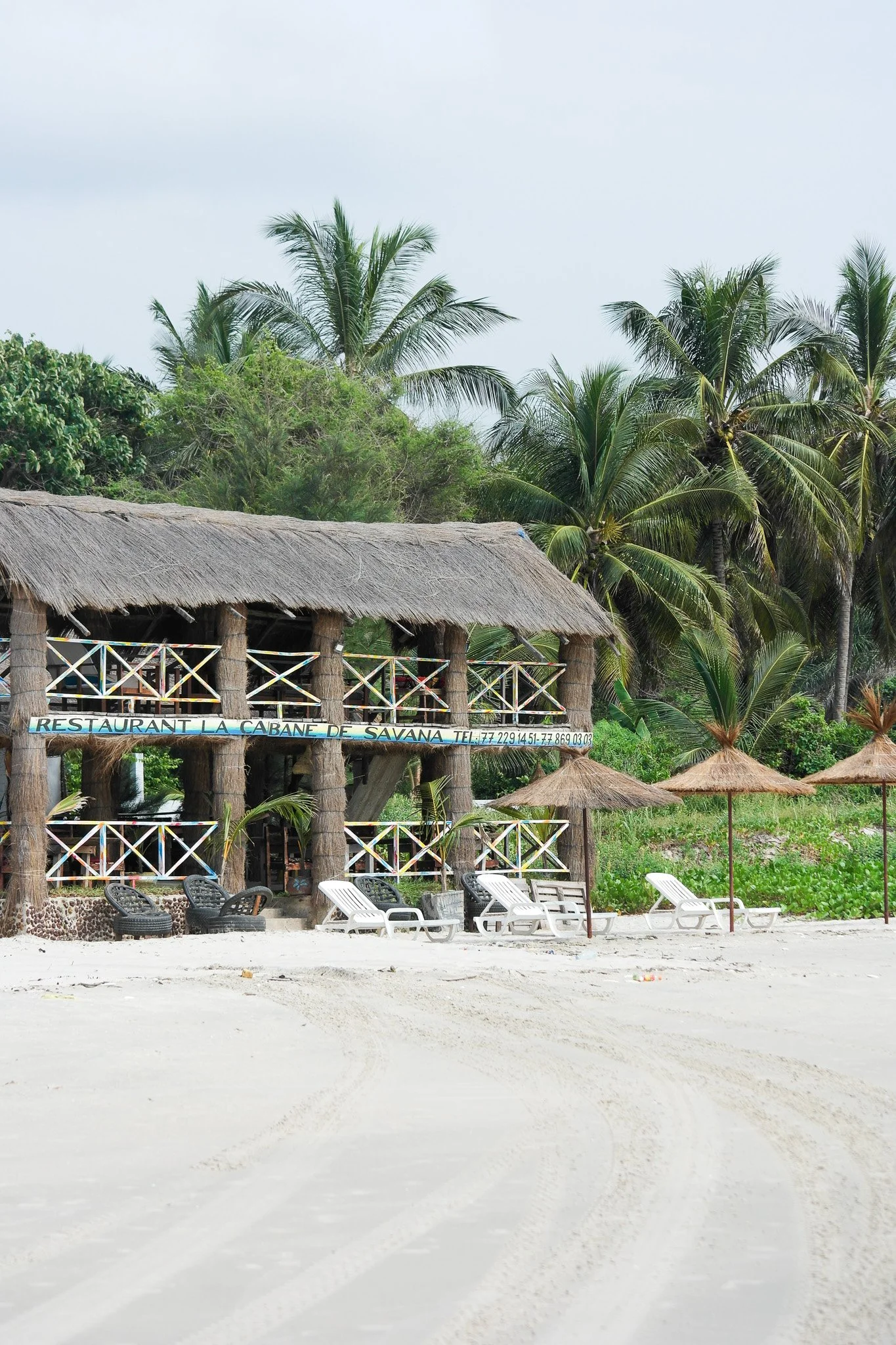 Une cabane en bois avec un toit en paille, située sur une plage de sable blanc, avec des chaises longues et des parasols en bambou, entourée de palmiers et d'une végétation tropicale.