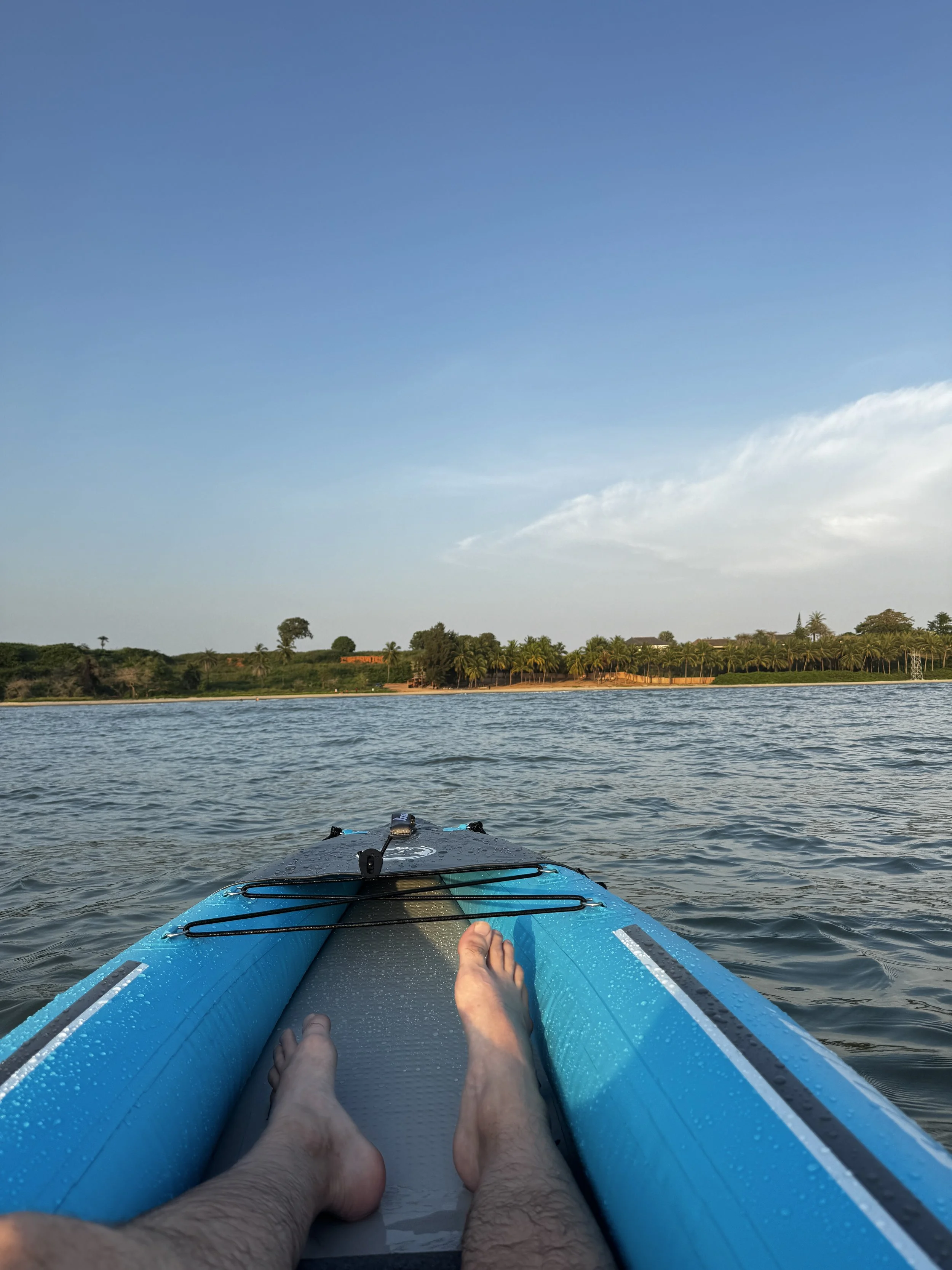 Personne allongée dans une planche de paddle sur un lac, avec une vue sur la rive avec des arbres et un ciel clair.