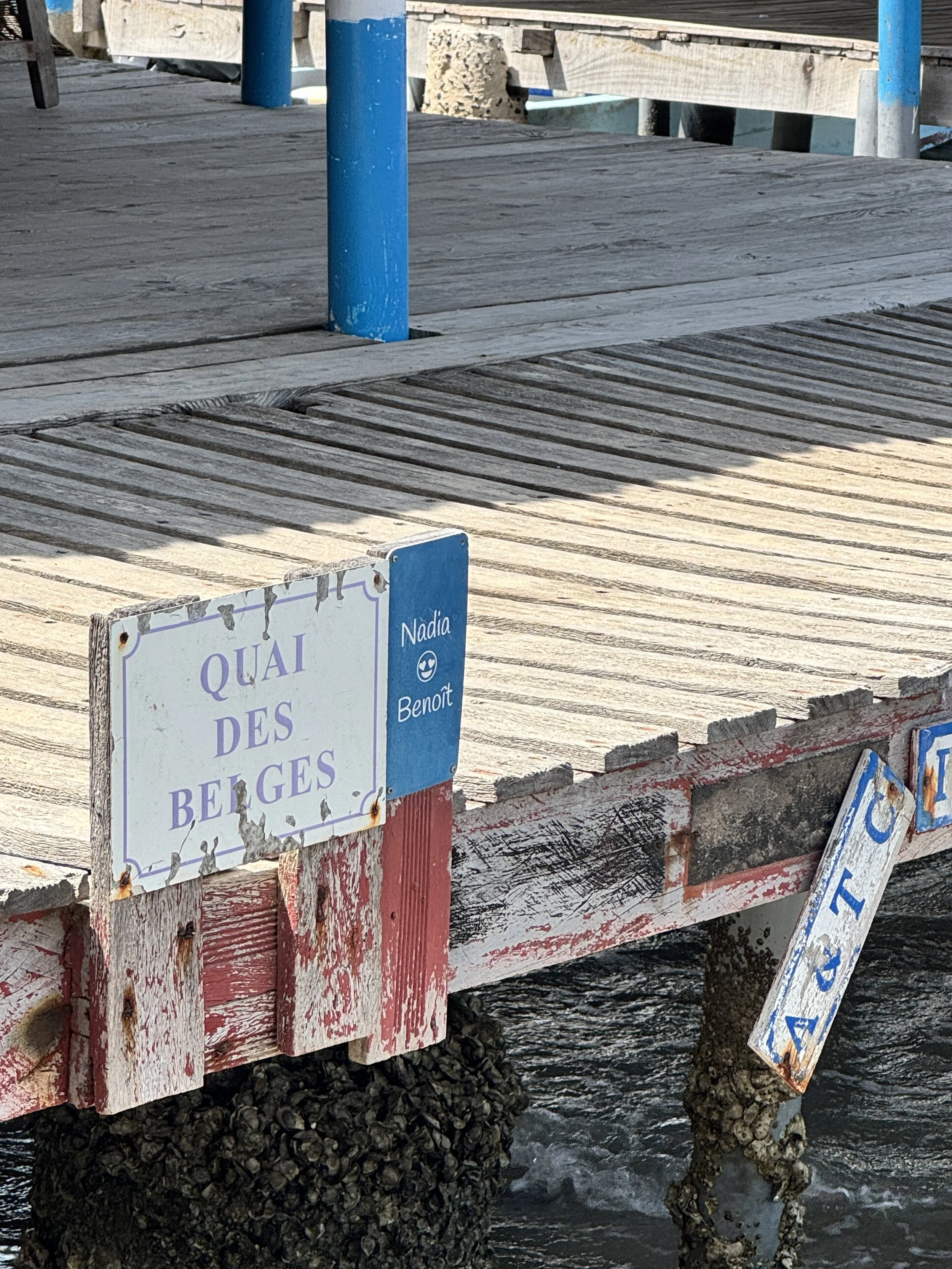 Pont en bois avec un panneau indiquant QUAI DES BELGES, à côté un panneau bleu avec le nom Nadia Benoît, et un autre panneau esseulé suspendu sous l'eau.