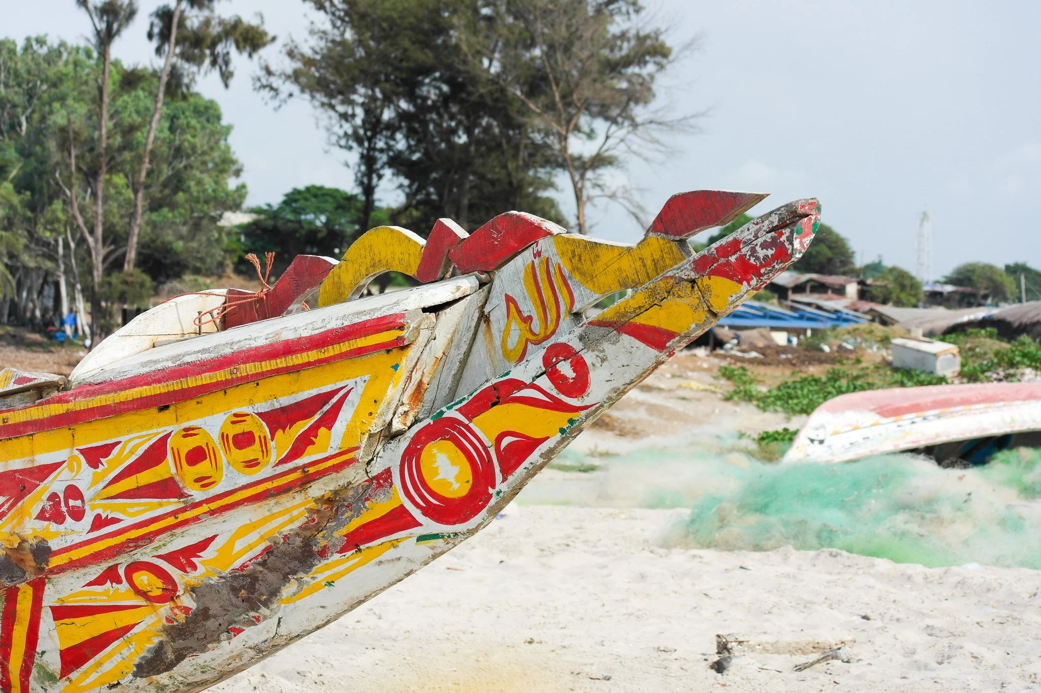 Un bateau en bois coloré et ancien sur une plage de sable, avec des arbres et des maisons en arrière-plan.