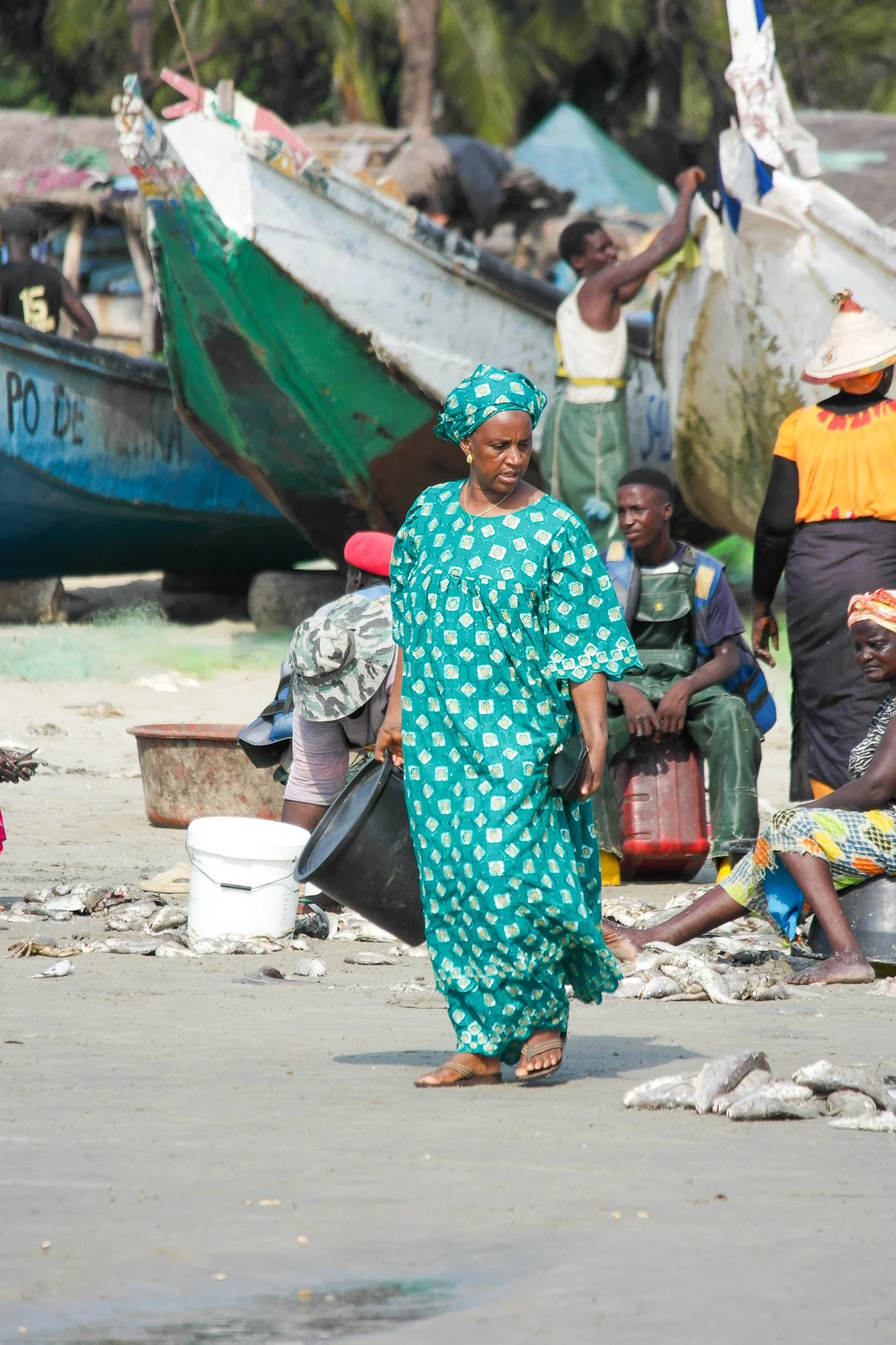 Femme en vêtement bleu à motifs portant un seau noir sur une plage, avec des pêcheurs et des bateaux en arrière-plan