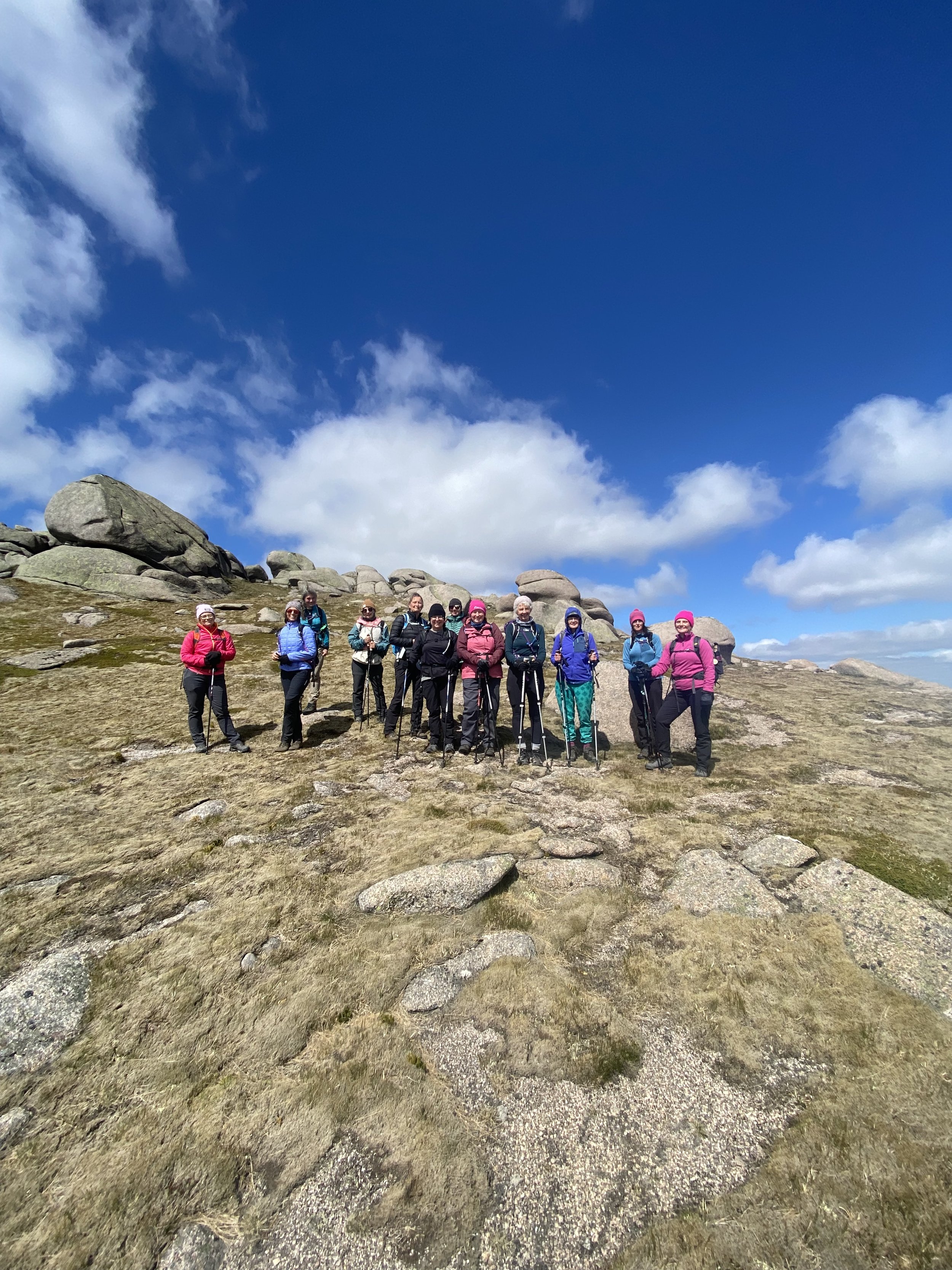 A group of hikers with backpacks and trekking poles standing on a rocky hillside under a partly cloudy blue sky.