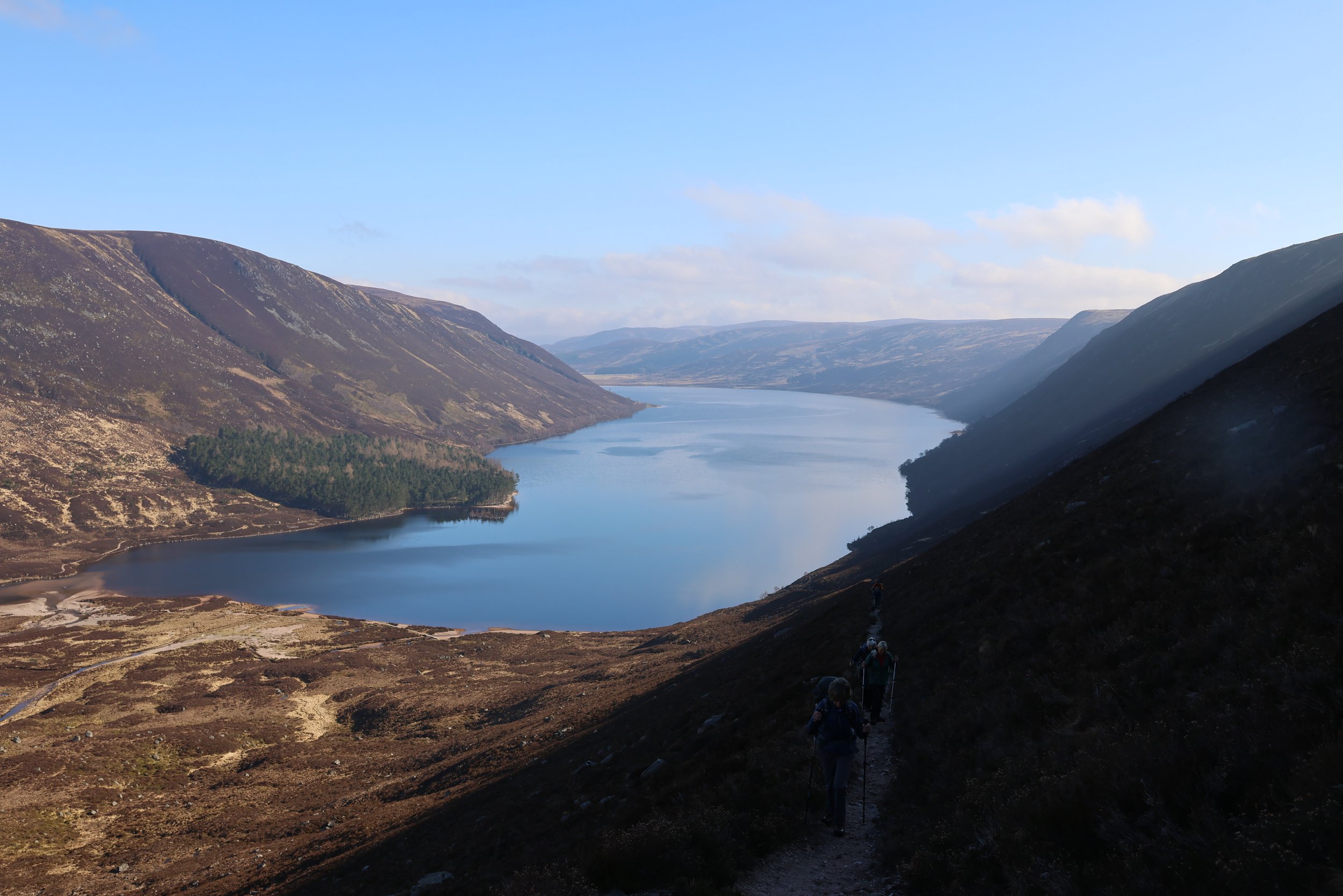 Hikers walking along a trail on a mountainside overlooking a large lake flanked by hills and mountains under a blue sky.