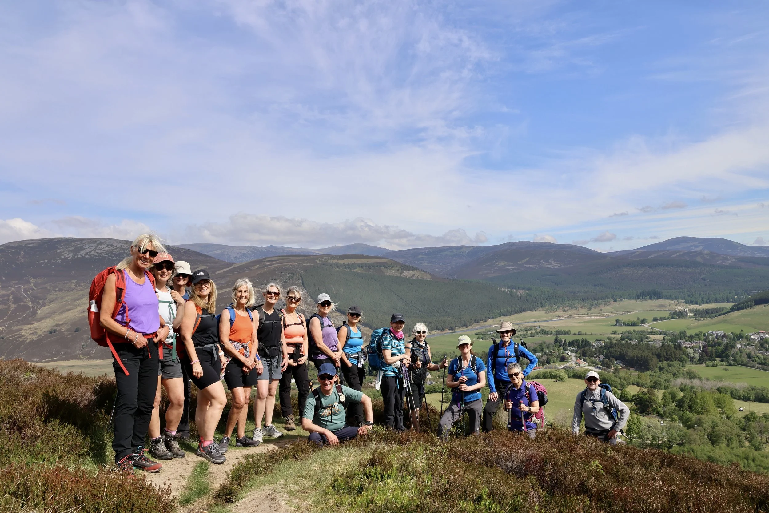 Group of hikers standing on a trail with mountains and green fields in the background under a partly cloudy sky.