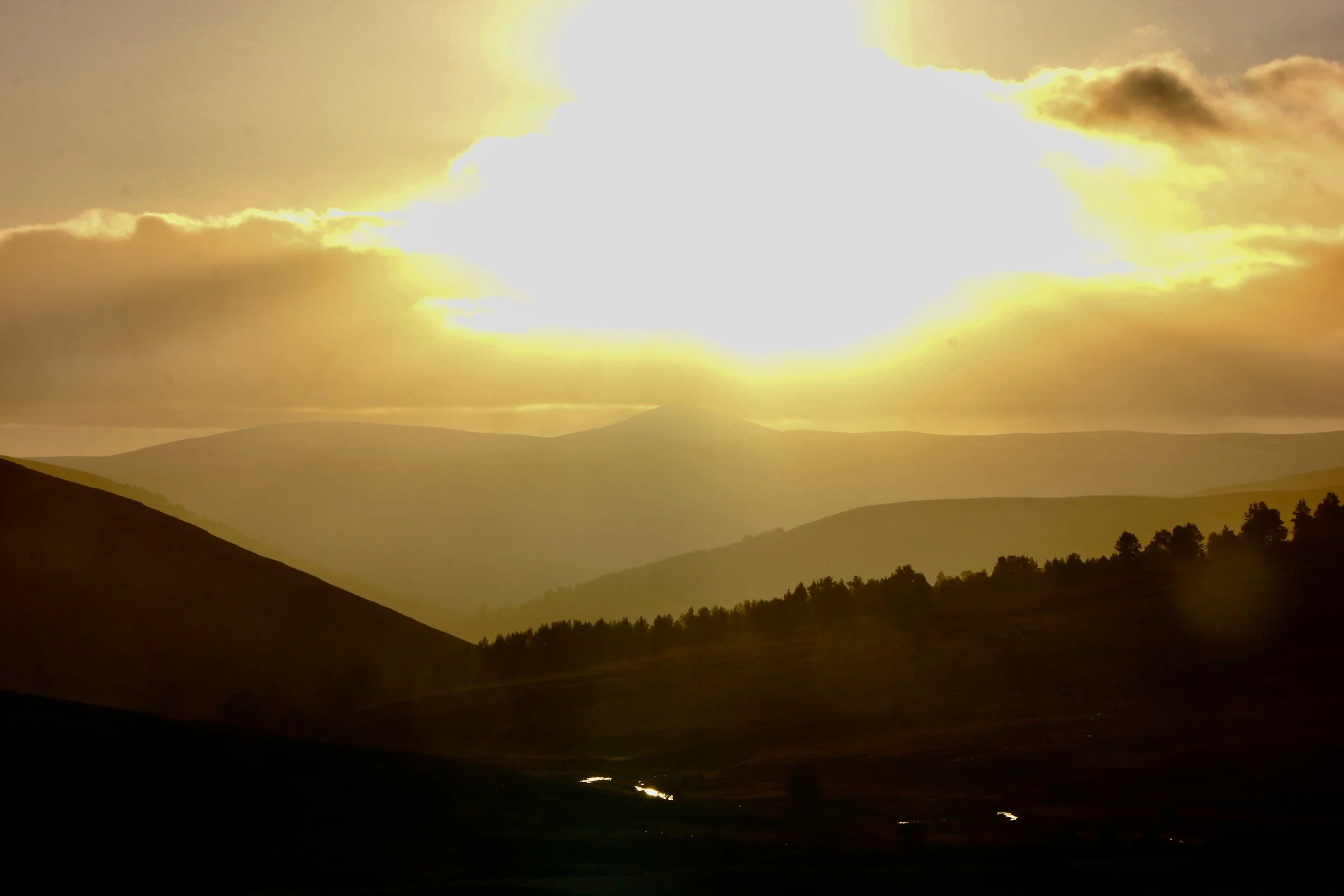 Sunset over rolling hills with a bright, partly cloudy sky and the sun near the horizon.