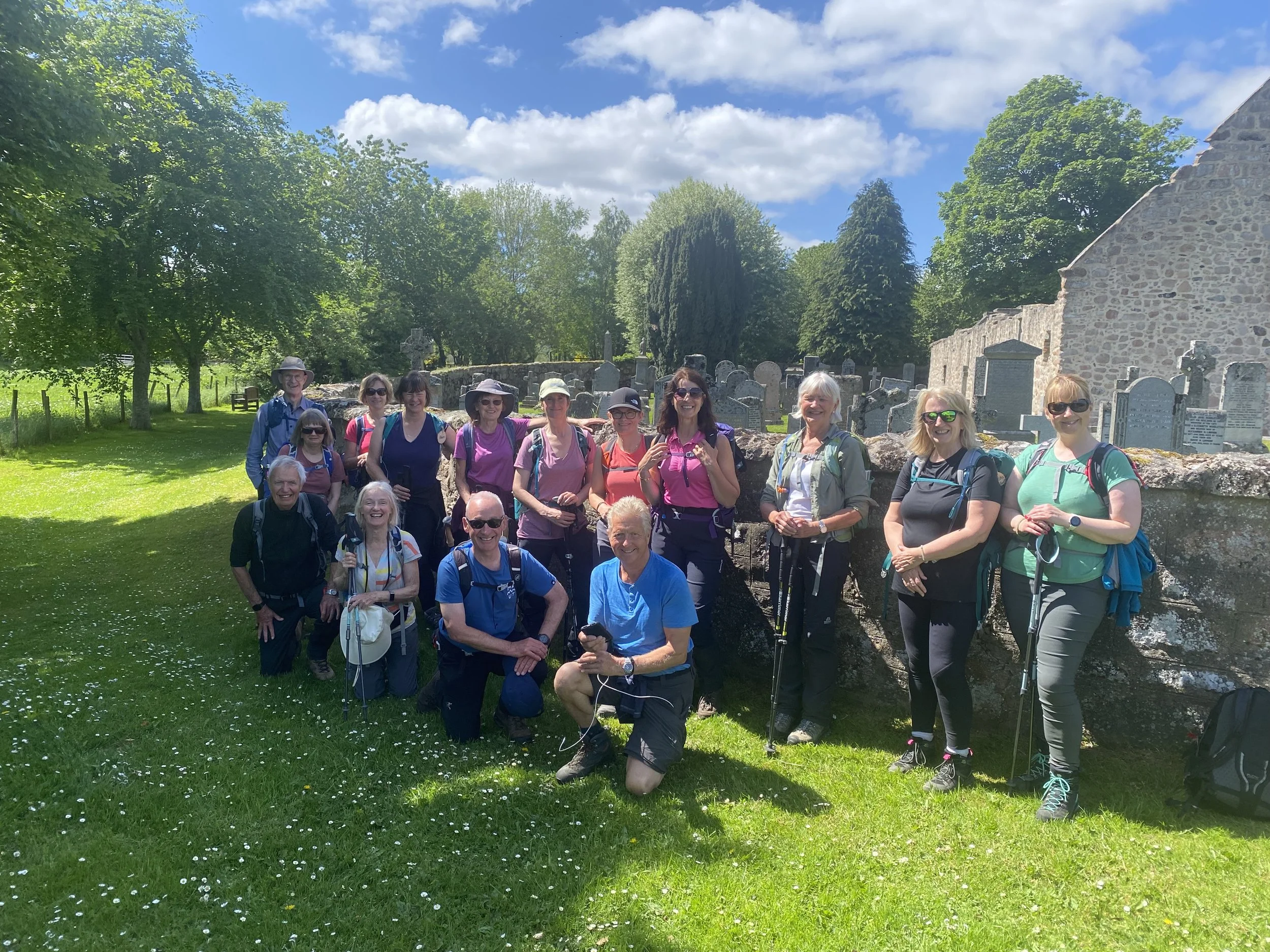 Group of 14 hikers, mostly women, standing on grassy area with trees and stone building ruins in background on a bright, sunny day.