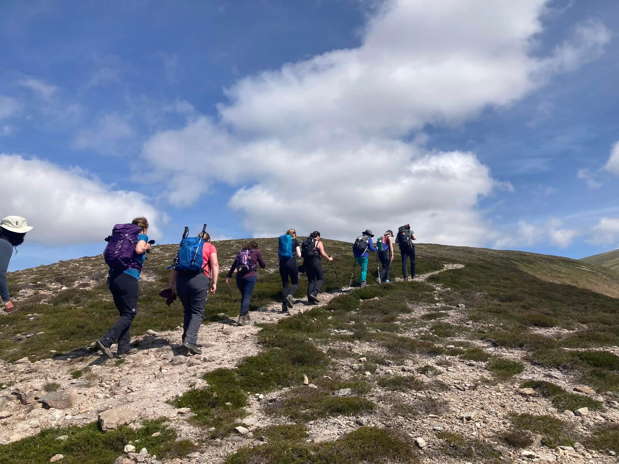 Group of hikers climbing a rocky trail on a mountain with green vegetation, under a partly cloudy blue sky.