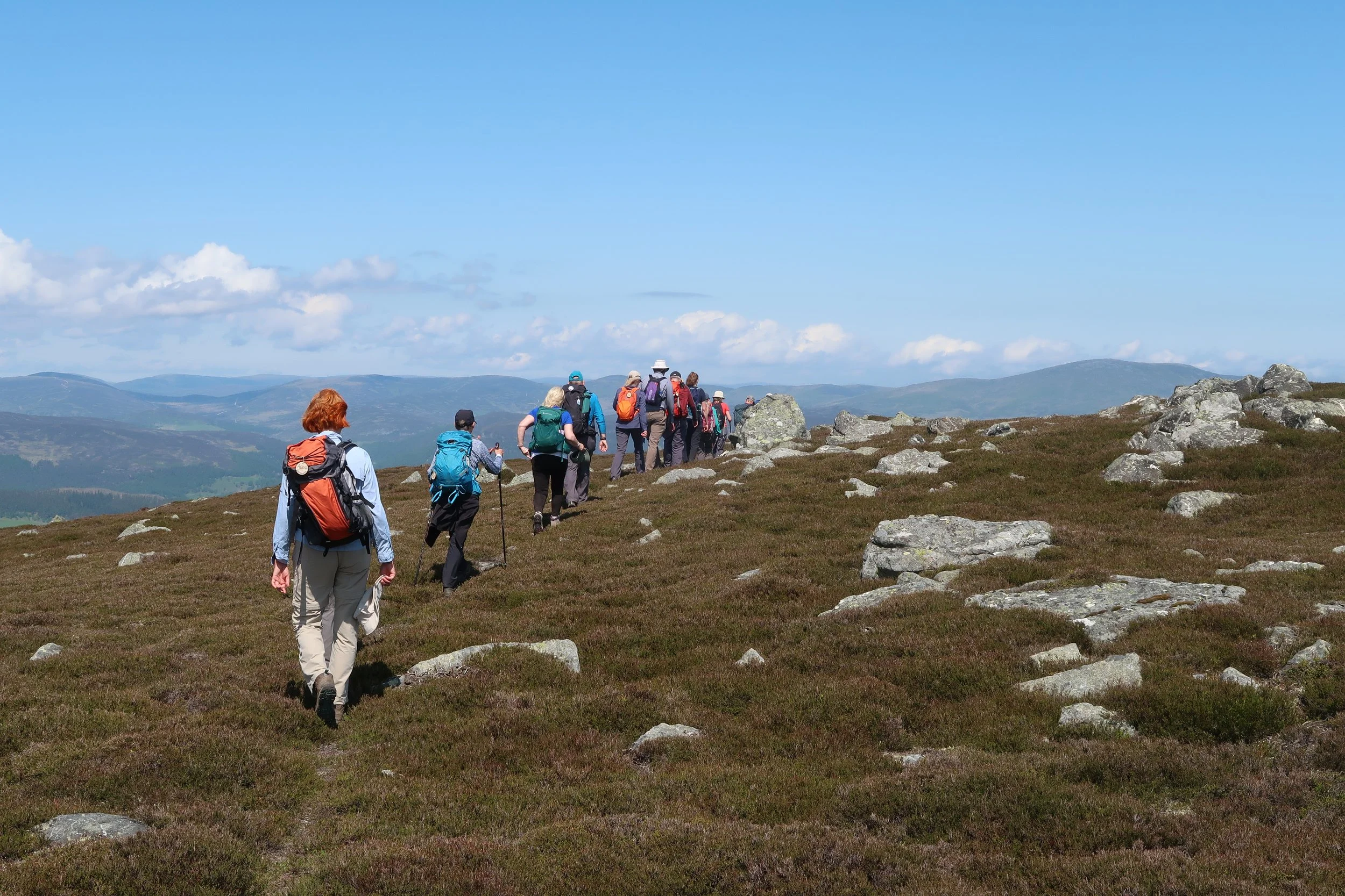 A group of hikers walking along a trail on a grassy hilltop with mountains in the background under a partly cloudy sky.