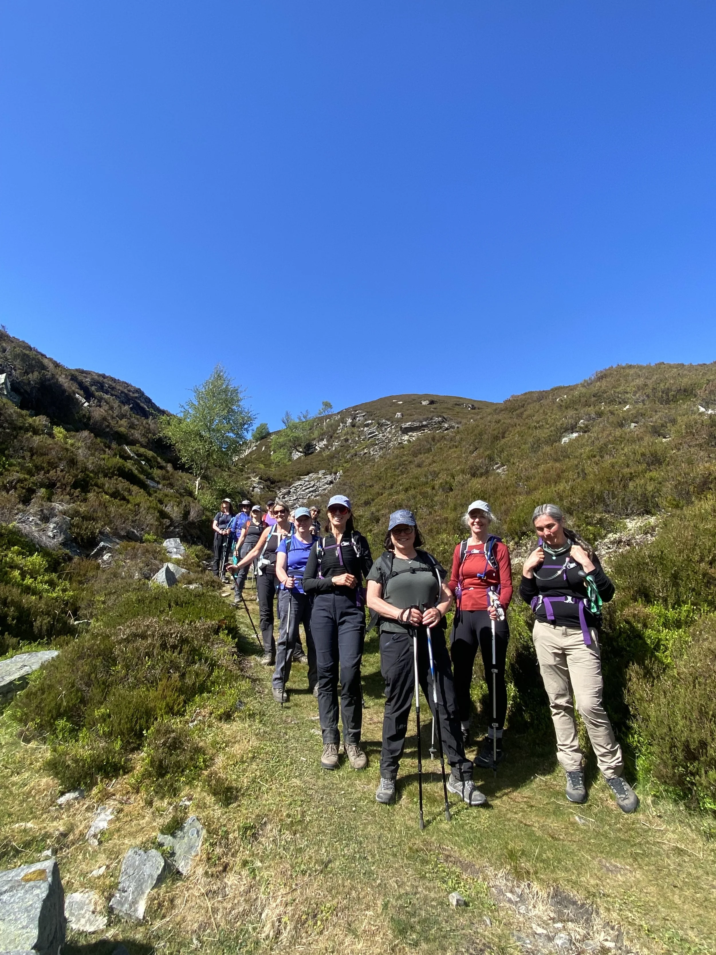 Group of eleven women hiking on a mountain trail under a clear blue sky, wearing outdoor clothing, hats, and holding trekking poles.