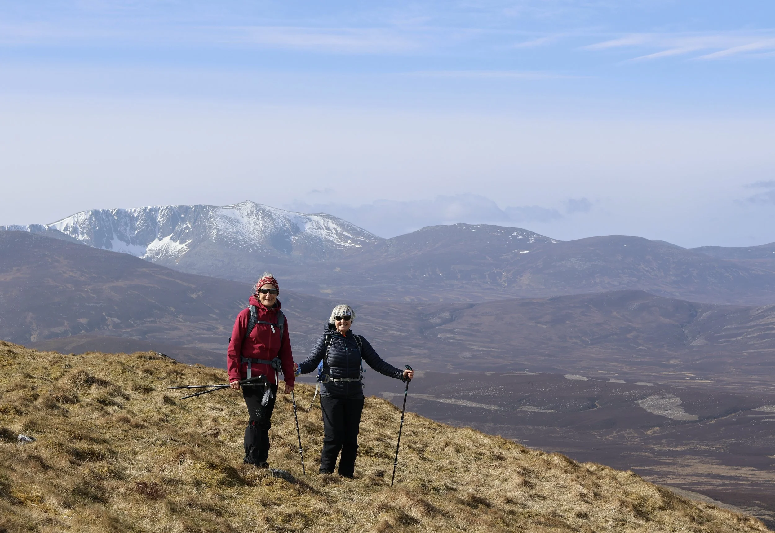 Two women hiking on a grassy mountain slope with snow-capped mountains in the background, wearing sunglasses and outdoor gear, holding hiking poles.