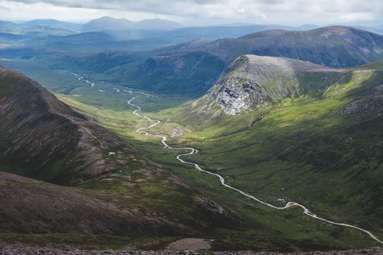 A scenic highland landscape with steep, green hills and a winding river flowing through a valley, under a partly cloudy sky.