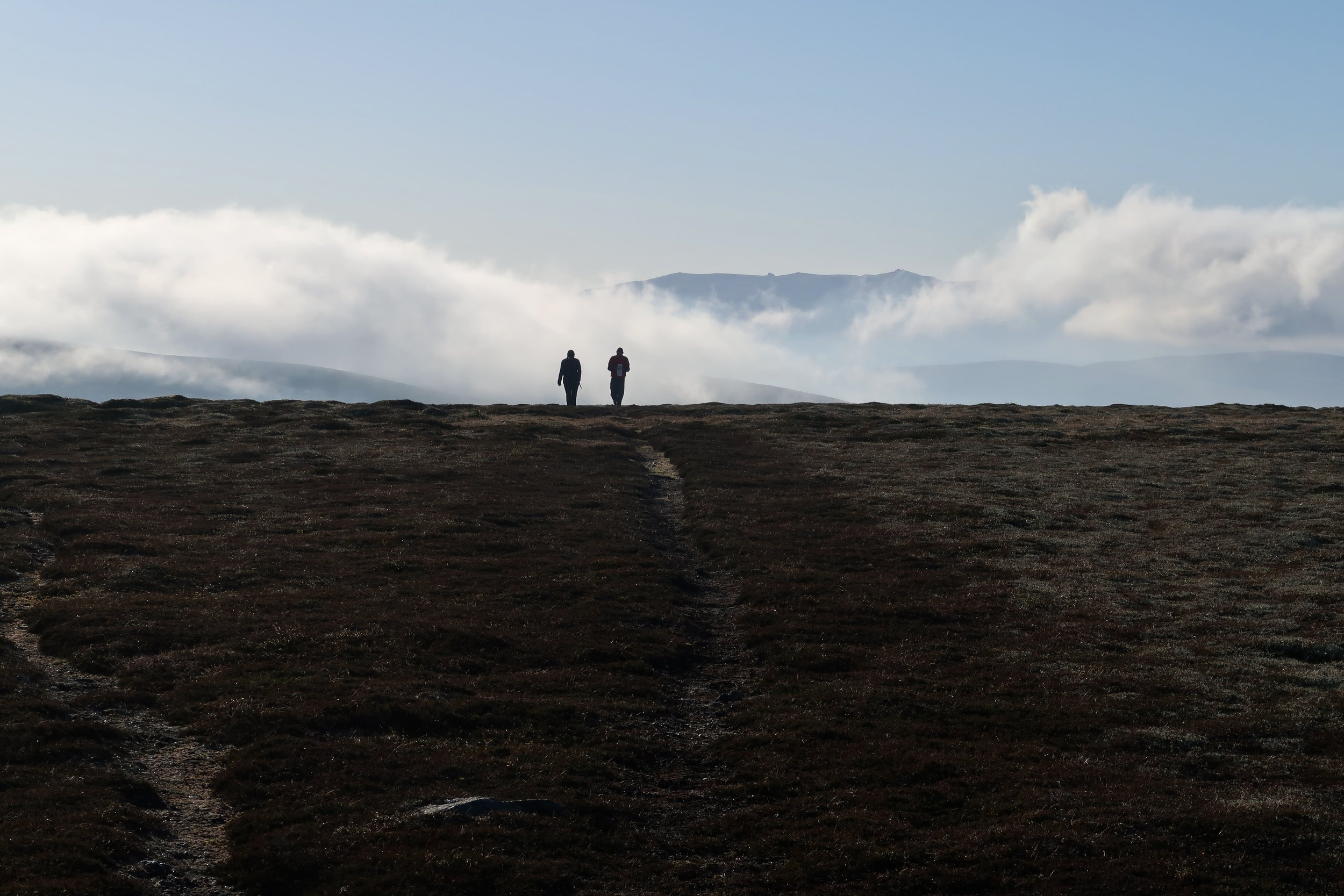 Two people walking on a dirt trail on a hilltop with a mountain range and clouds in the background.